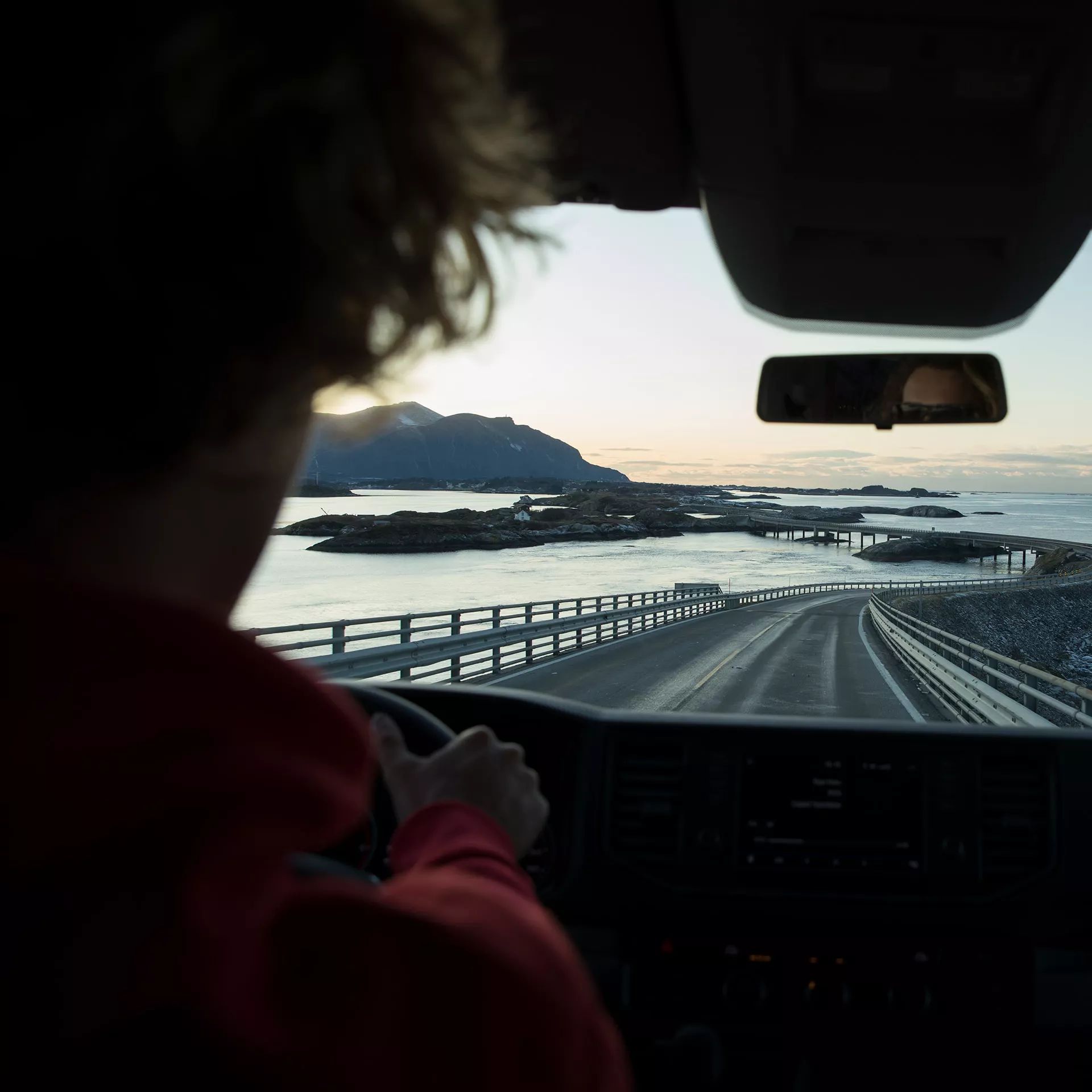 Eine Person fährt auf einer Küstenstraße bei Sonnenuntergang. Die Straße schlängelt sich vorbei an Wasser und Bergen. Der Blick aus dem VW Grand California zeigt teils bedeckte Hintergründe und ein spiegelndes Meer.