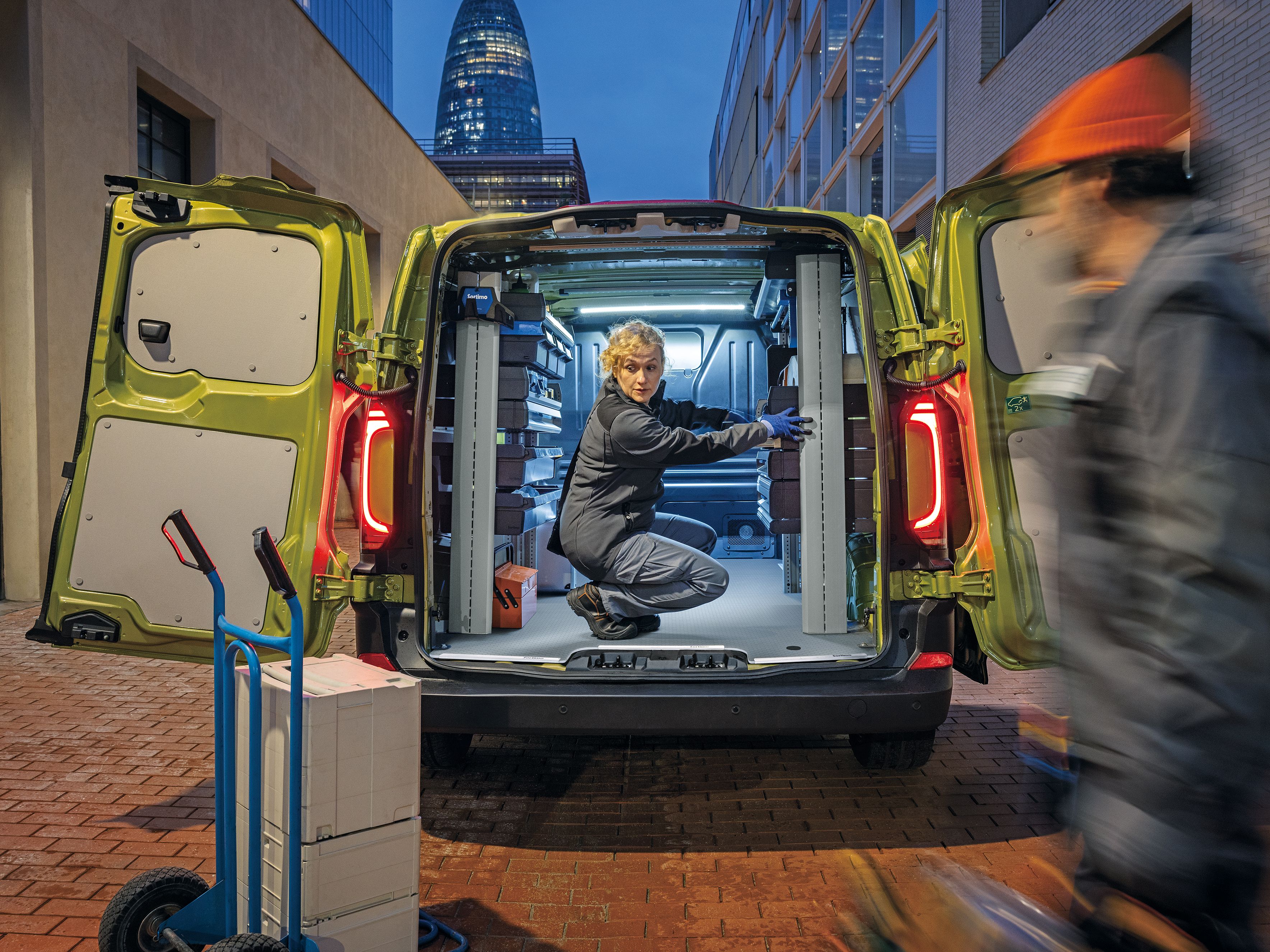 Жена организира работно оборудване във Volkswagen Transporter с отворени задни врати на градска улица