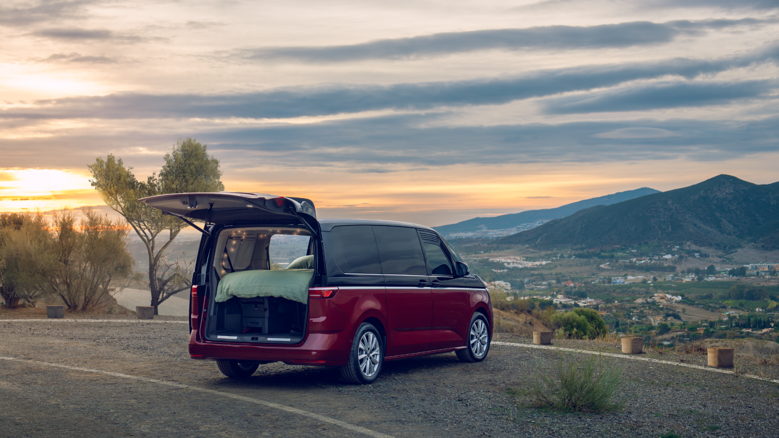 Volkswagen Multivan с отворен заден багажник, превърнат в уютно място за спане, паркиран сред живописен пейзаж по залез