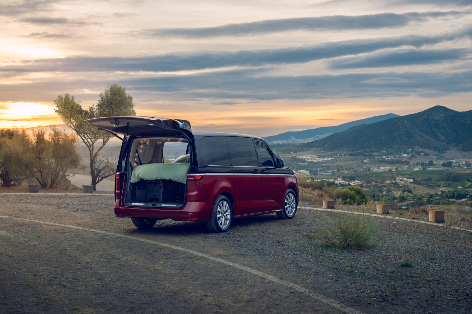 Volkswagen Multivan с отворен заден багажник, превърнат в уютно място за спане, паркиран сред живописен пейзаж по залез