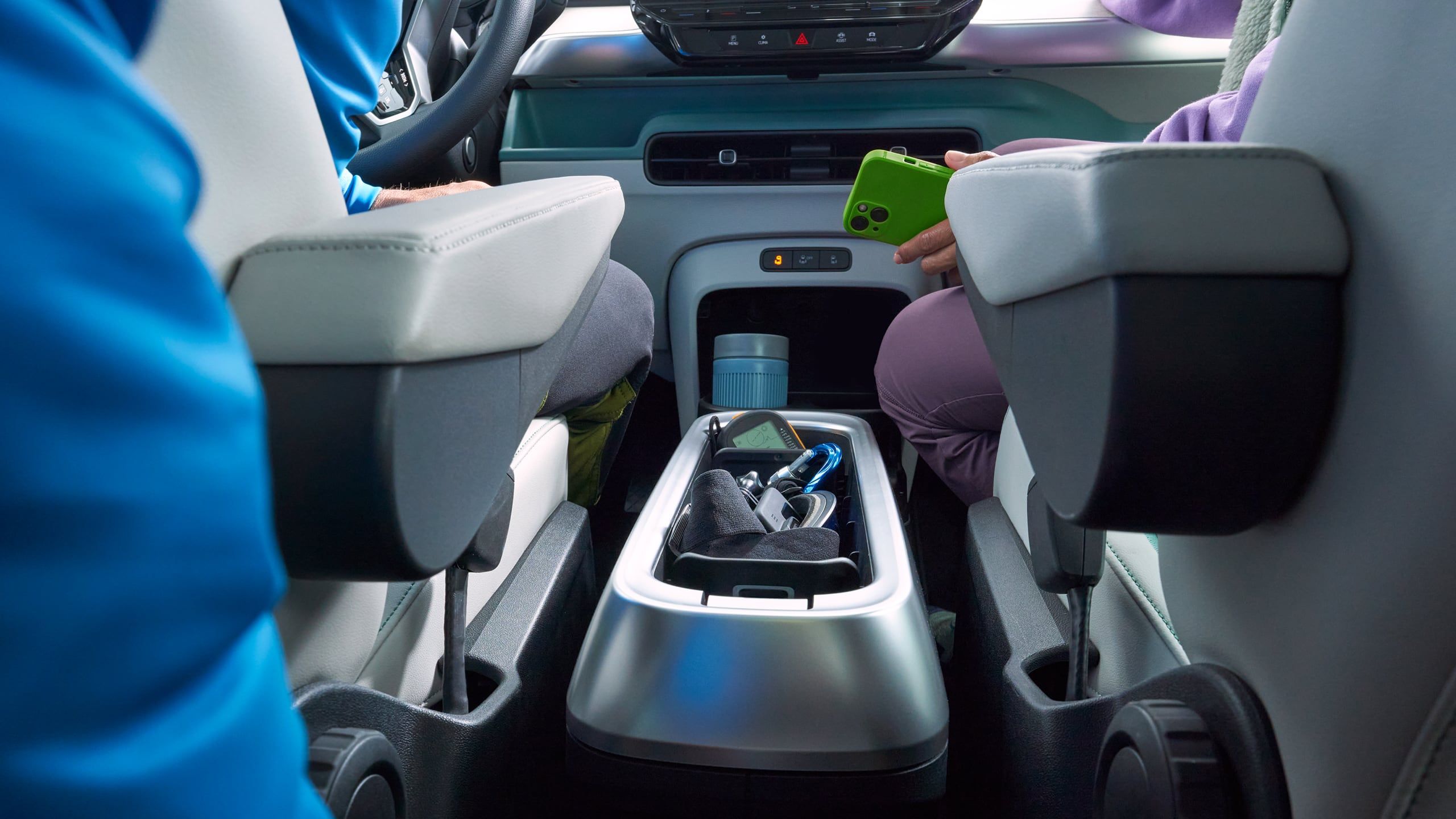 Поглед към централната конзола на автомобил, включваща бутилка за вода и мобилен телефон. На предните седалки са заети от двама души, единият от които държи зелен телефон.