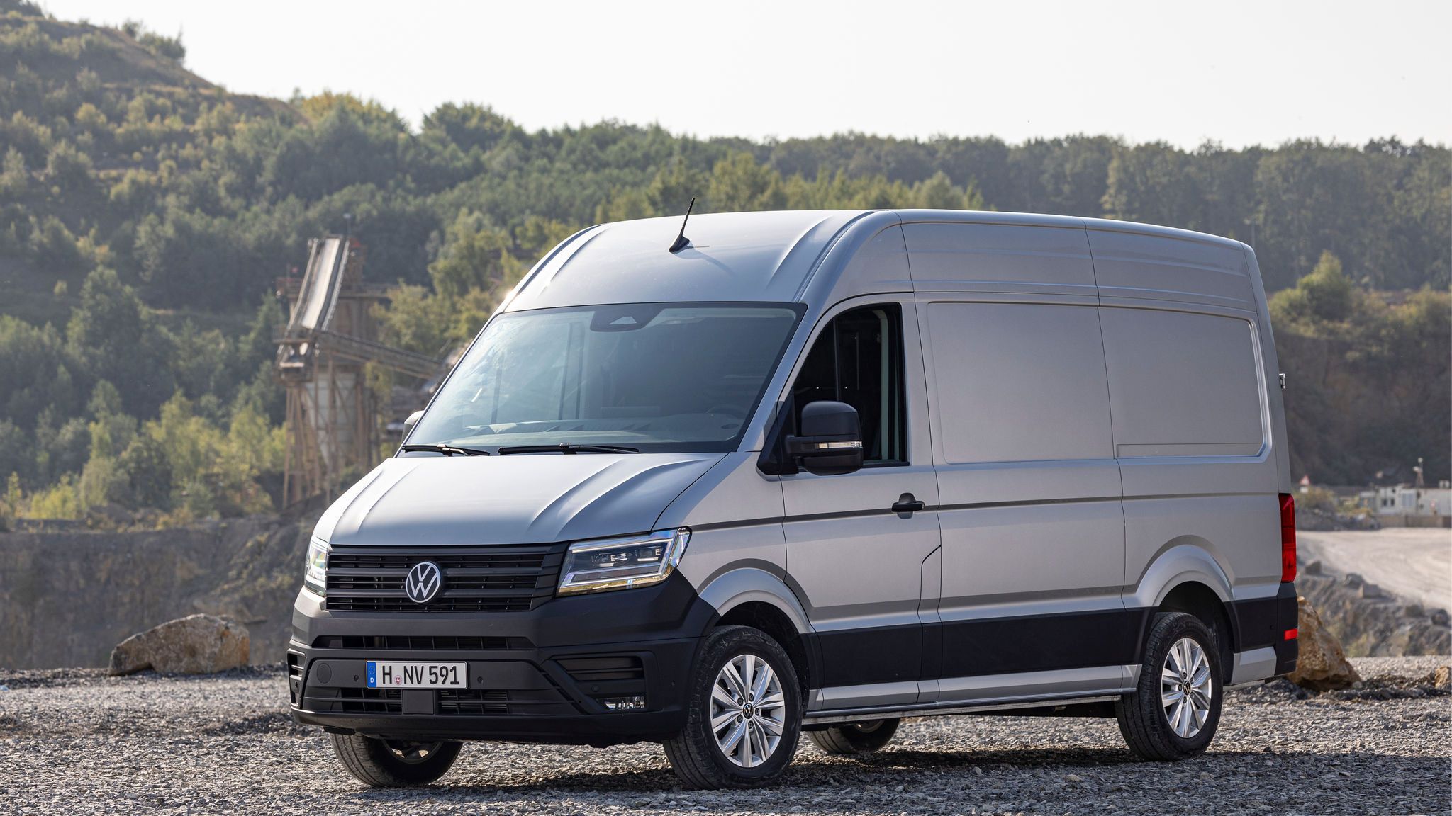 Сребрист Volkswagen Crafter, паркиран на чакъл в индустриален район с горски фон.