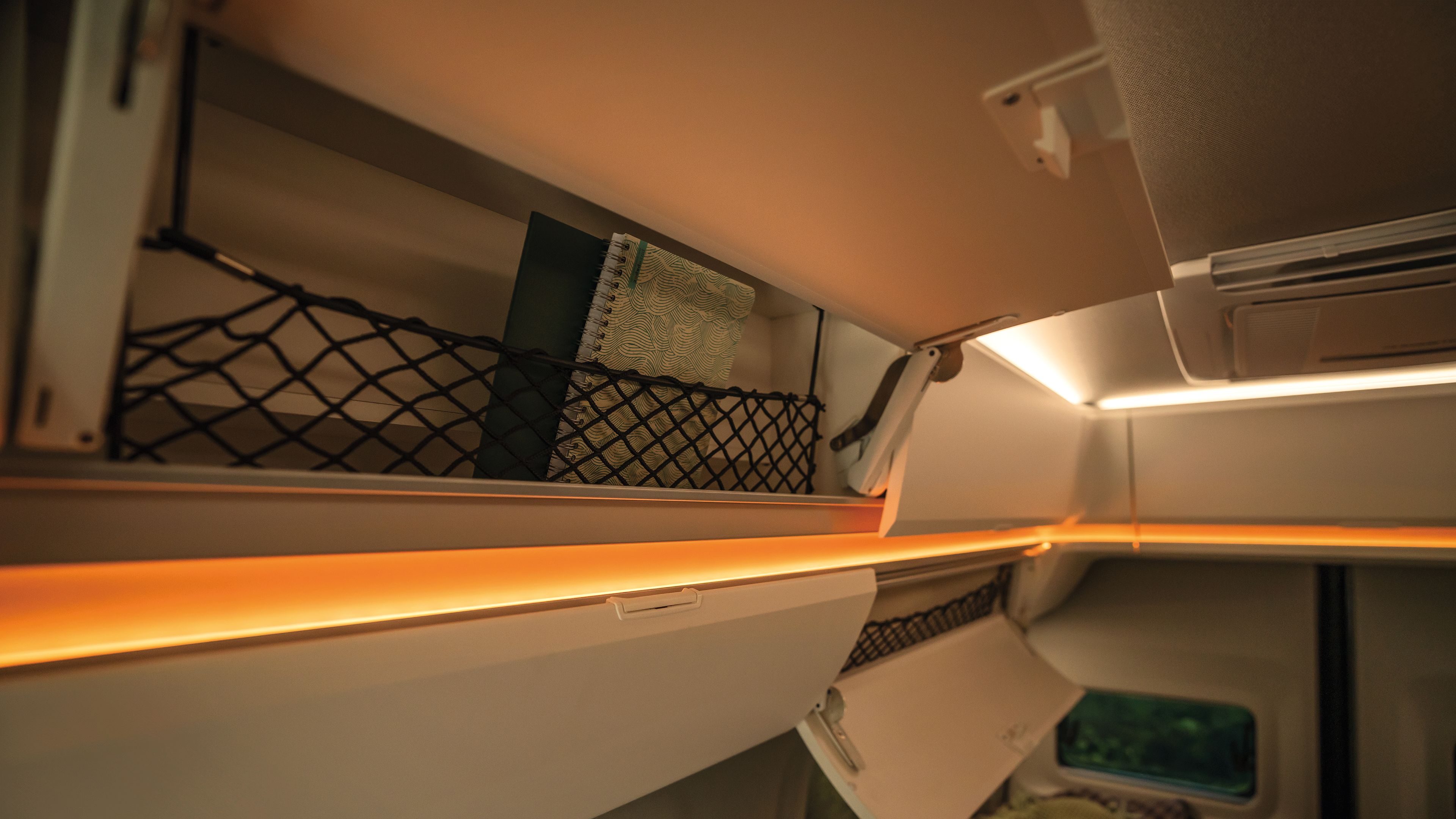 Darstellung des Innenraums eines Wohnmobils mit einer Netzablage, in der sich Bücher befinden. Es gibt Beleuchtung entlang der Kanten und ein Fenster ist teilweise im Bild sichtbar. Ein Bett mit Kissen ist ebenfalls zu sehen.