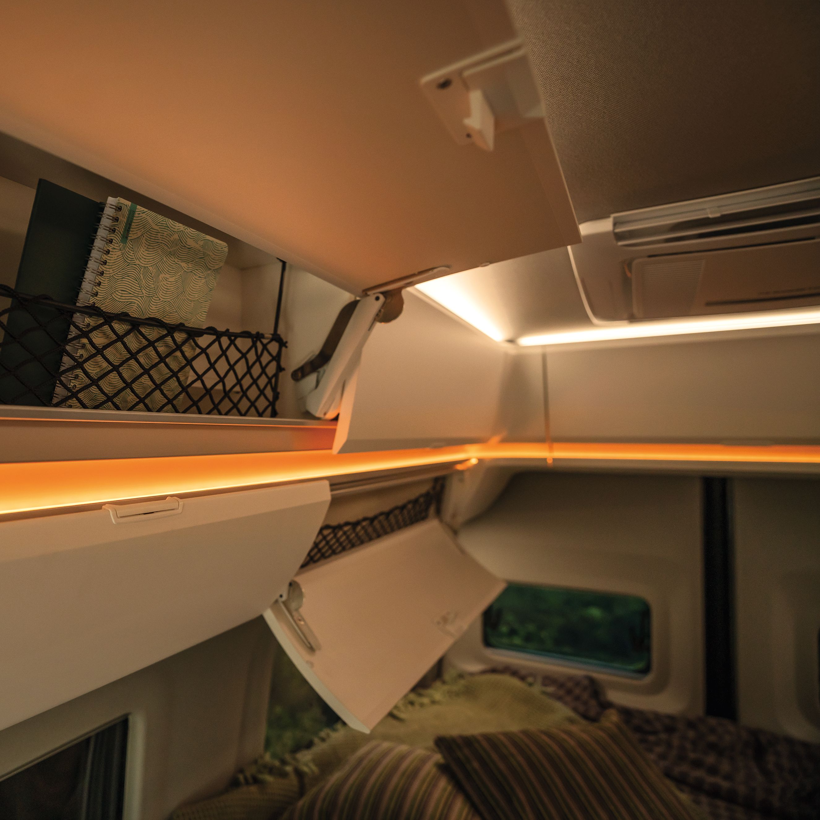 Darstellung des Innenraums eines Wohnmobils mit einer Netzablage, in der sich Bücher befinden. Es gibt Beleuchtung entlang der Kanten und ein Fenster ist teilweise im Bild sichtbar. Ein Bett mit Kissen ist ebenfalls zu sehen.