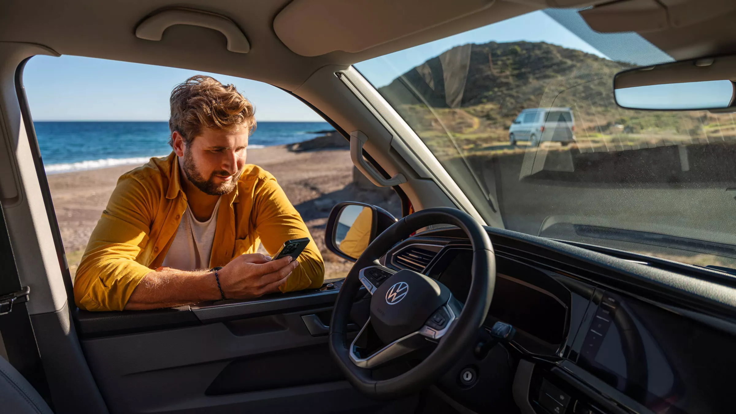 Мъж до Volkswagen Multivan на плажа, гледащ телефона си през отворения прозорец.
