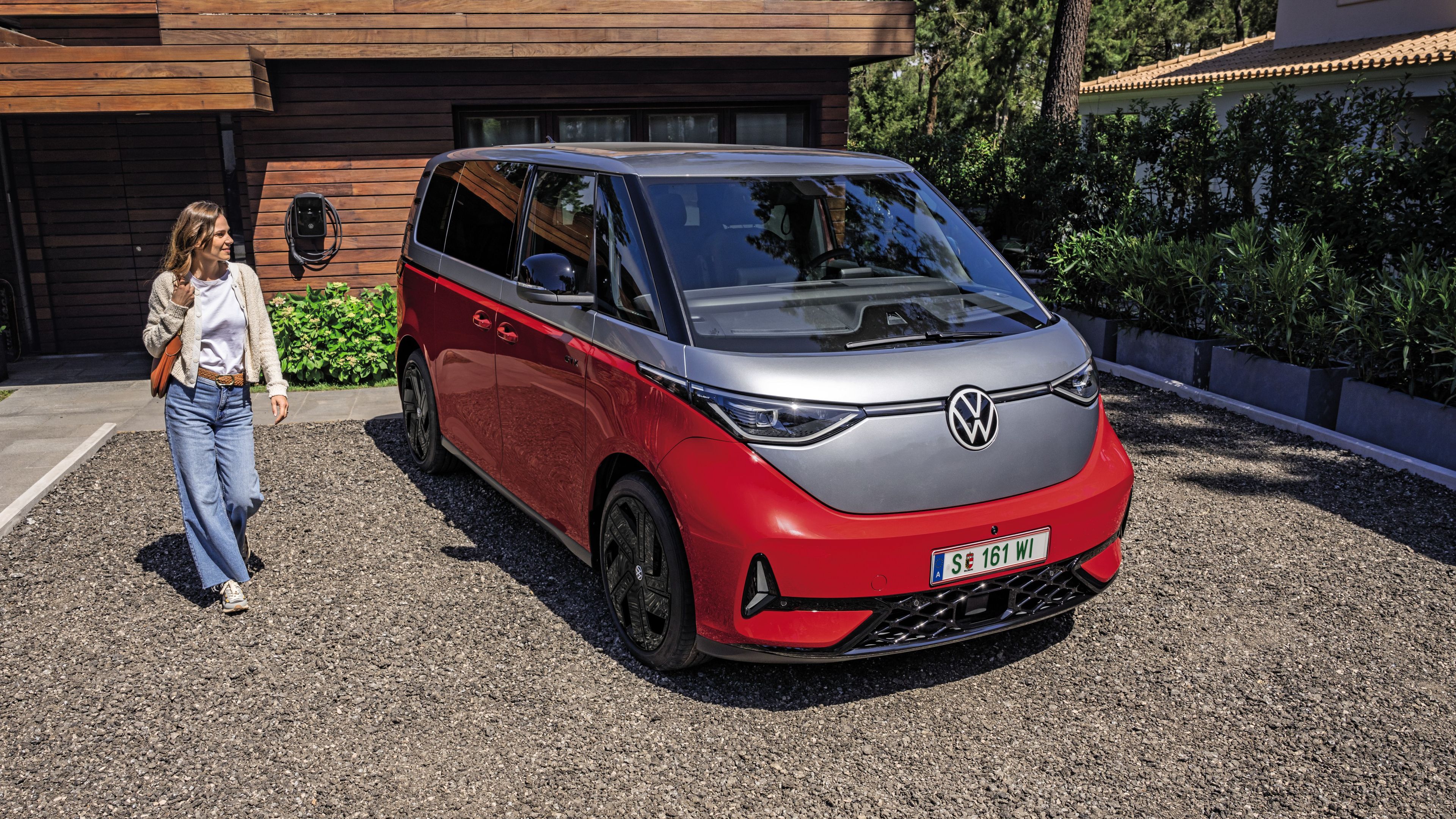 Жена се приближава към червено-сив Volkswagen ID. Buzz, паркиран пред модерен дом с инсталирана зарядна станция.