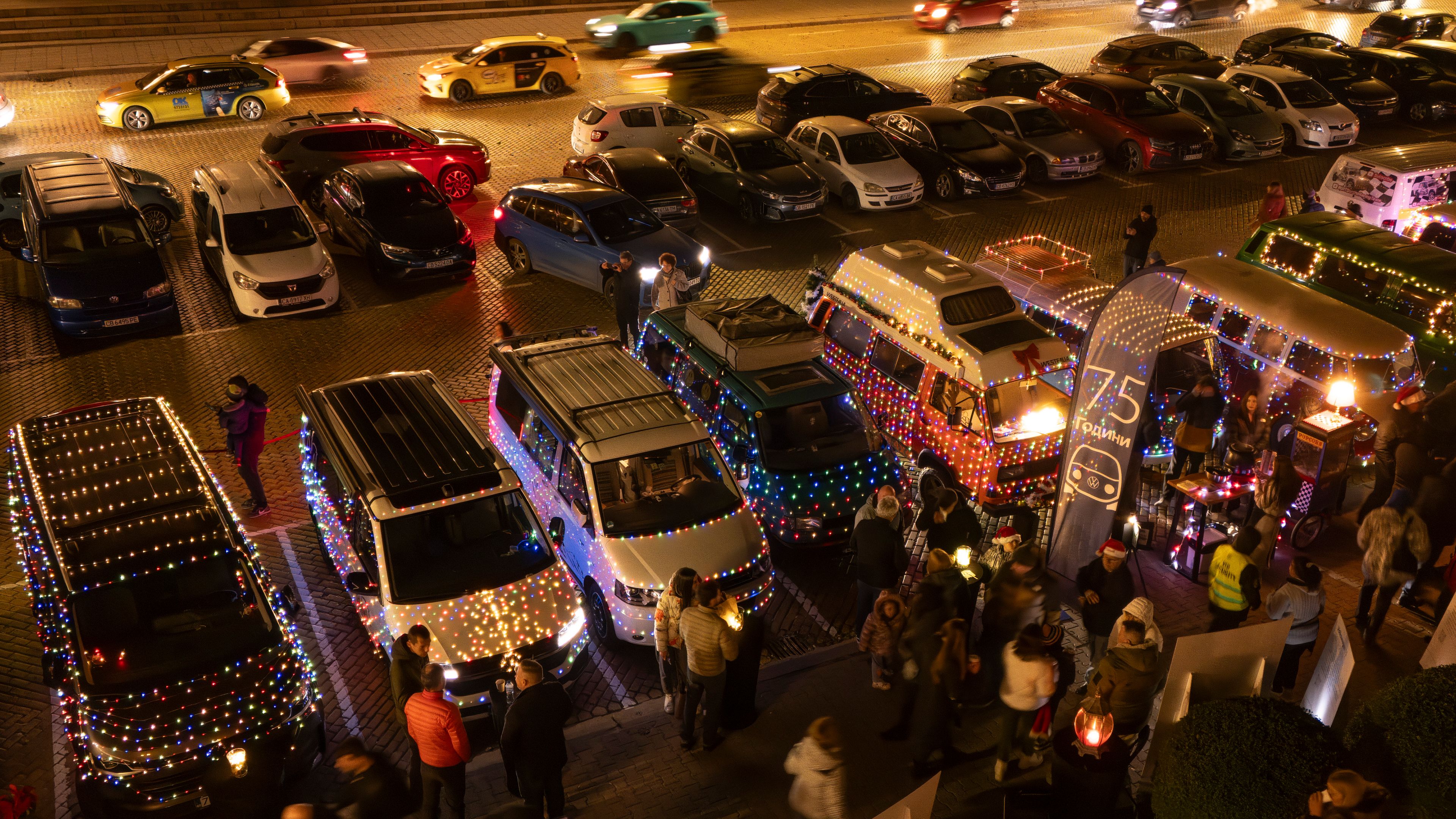 Автомобили Volkswagen, украсени със светлинни гирлянди, паркирани на оживен паркинг през нощта. Пред тях има група хора, които се разхождат и наблюдават осветените коли. На заден план се виждат преминаващи автомобили.