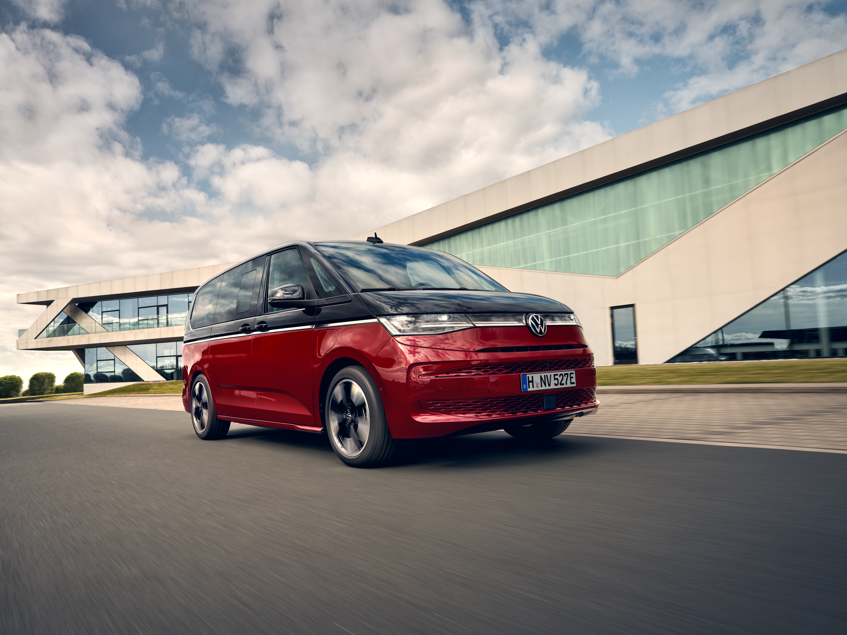 Volkswagen Multivan в движение пред съвременна архитектура. Червен Volkswagen Multivan се движи по път пред модерна сграда.