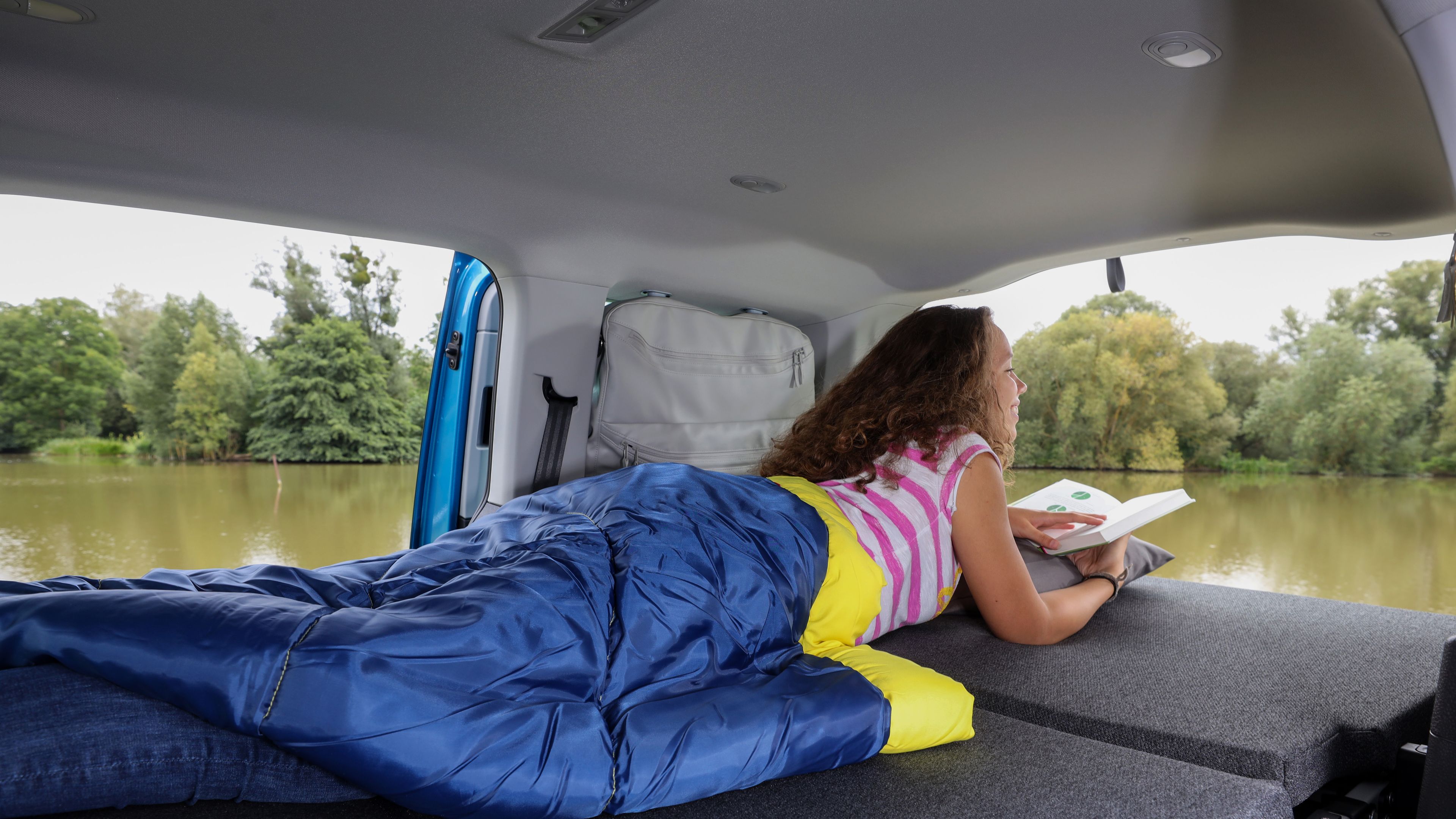 Frau liegt in Bett von VW Caddy California und liest ein Buch.