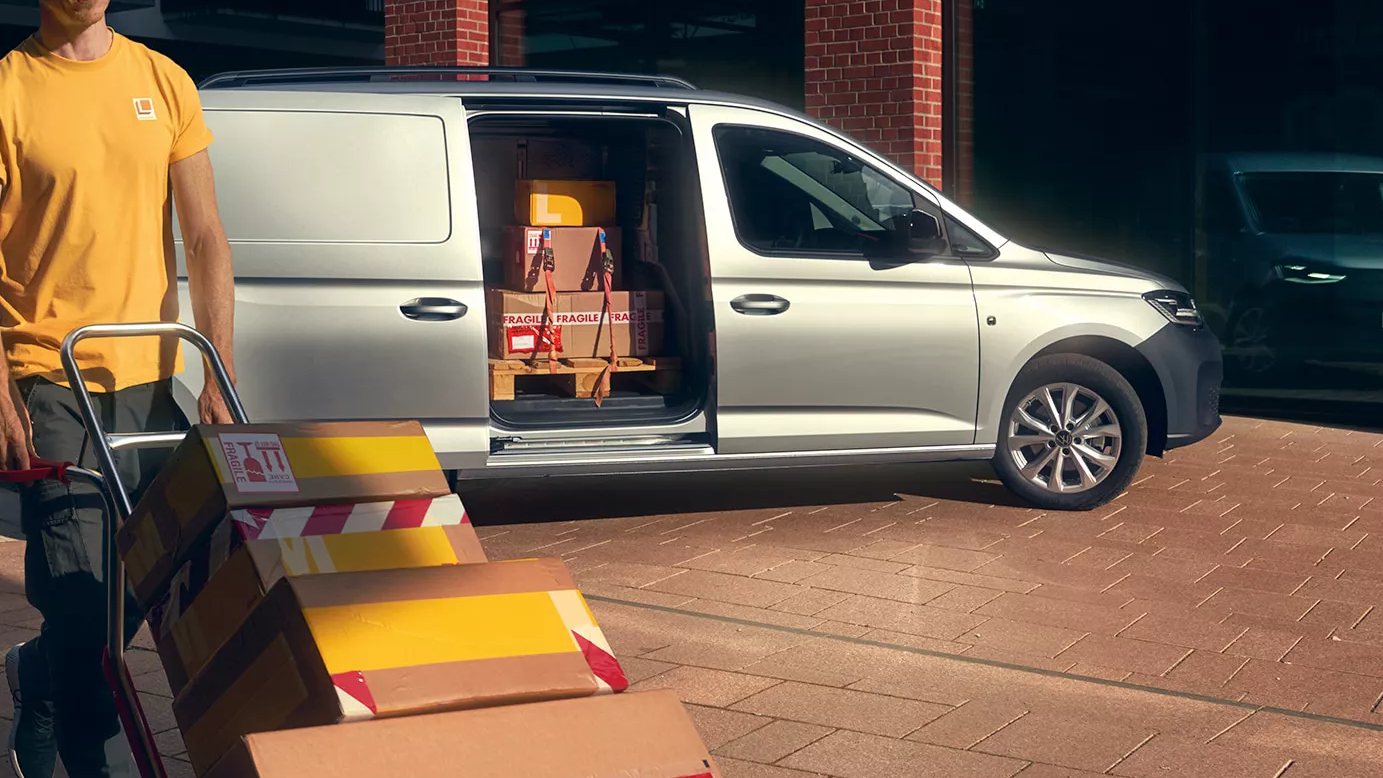 Куриер с количка пренася пакети пред сребрист товарен автомобил Volkswagen с отворена странична врата, натоварен с кашони.