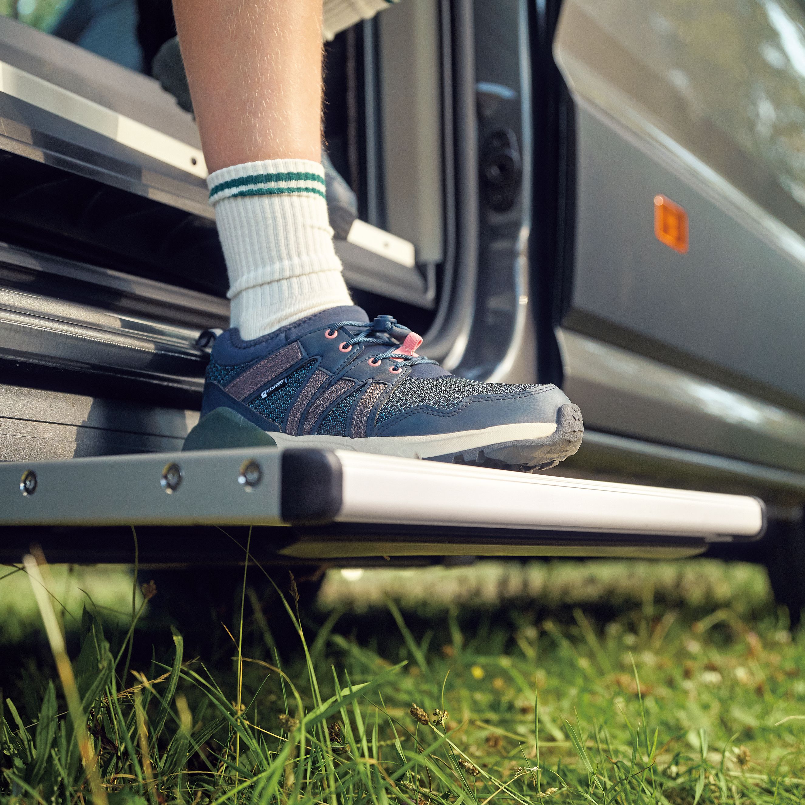 Eine Person mit weißen Socken und blauen Sneakern steht auf der ausziehbaren Trittstufe eines Campingwagens. Der Campingwagen steht in einer grünen Umgebung mit Gras. Die Trittstufe ist teilweise ausgefahren und ermöglicht einen bequemen Einstieg.