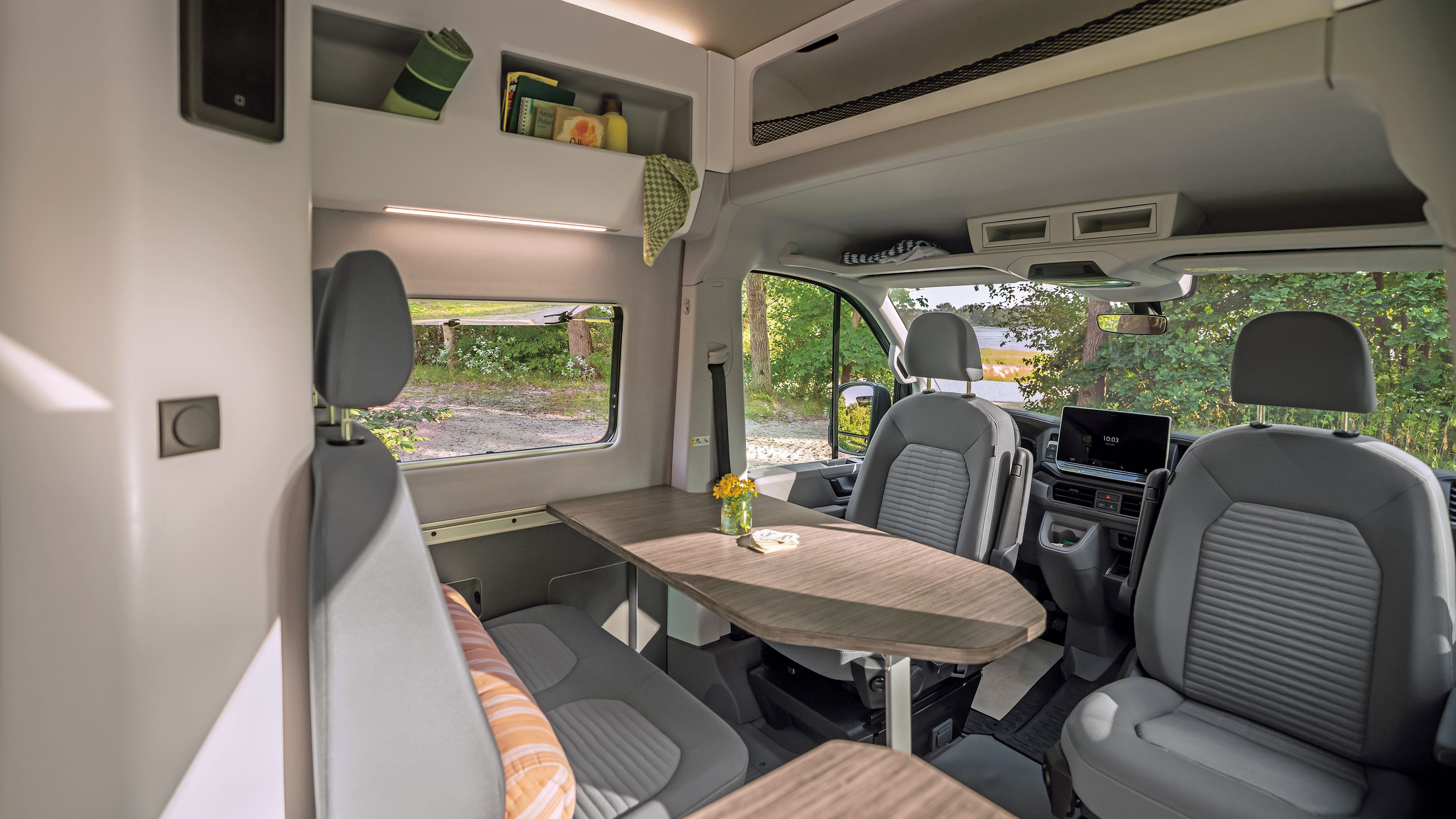 Das Bild zeigt das Innere eines Wohnmobils mit zwei grauen Vordersitzen, einem Tisch und einer kleinen Vase mit Blumen darauf. Im Hintergrund sind Regale mit ein paar Gegenständen zu sehen. Durch das Fenster ist ein grüner Außenbereich sichtbar.