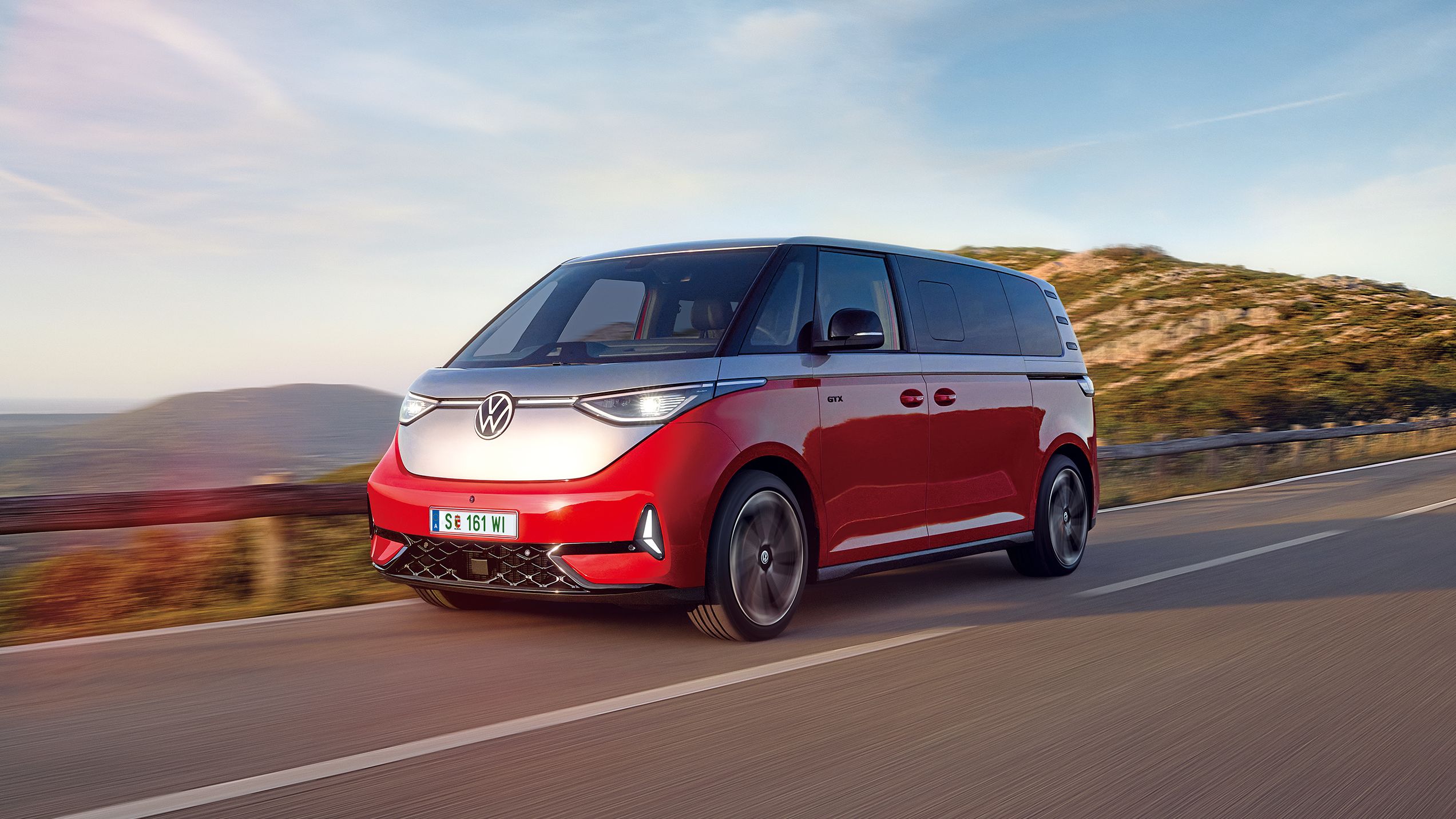  Volkswagen ID. Buzz GTX в червено-сив цвят в движение по живописен планински път при залез.