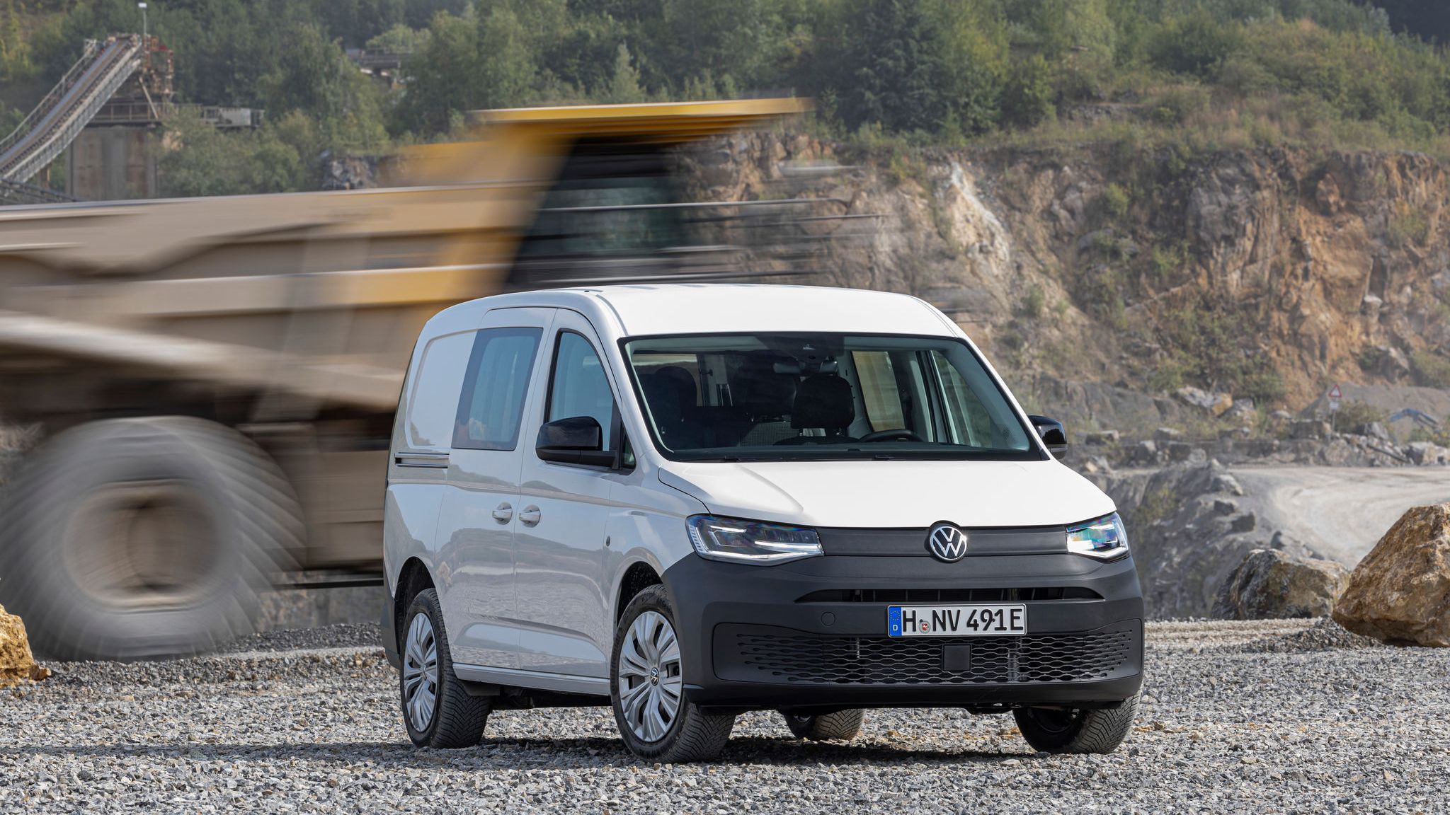 Бял Volkswagen Caddy Cargo паркиран на чакъл с преминаващ тежкотоварен камион на заден план