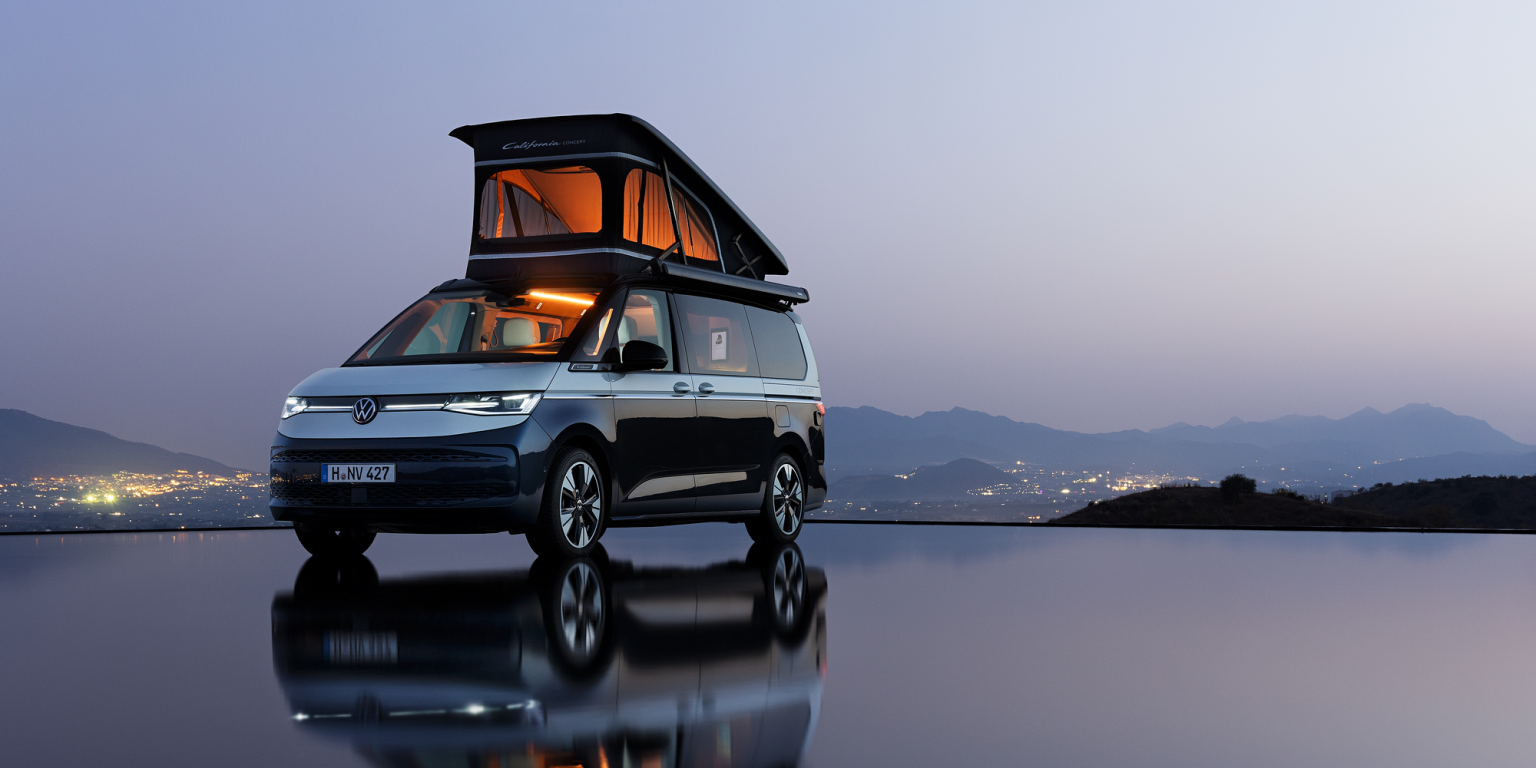 Volkswagen California – дом на колела под звездите Кемпер Volkswagen California с повдигнат покрив, паркиран край воден басейн с планински силуети и градски светлини на хоризонта.