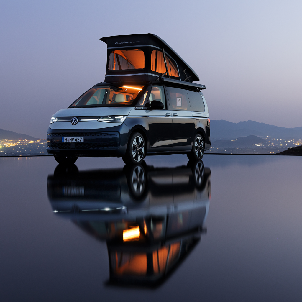 Кемпер Volkswagen California с повдигнат покрив, паркиран край воден басейн с планински силуети и градски светлини на хоризонта.