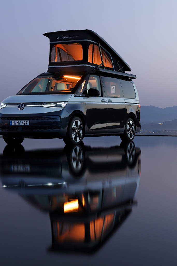 Volkswagen California – дом на колела под звездите Кемпер Volkswagen California с повдигнат покрив, паркиран край воден басейн с планински силуети и градски светлини на хоризонта.