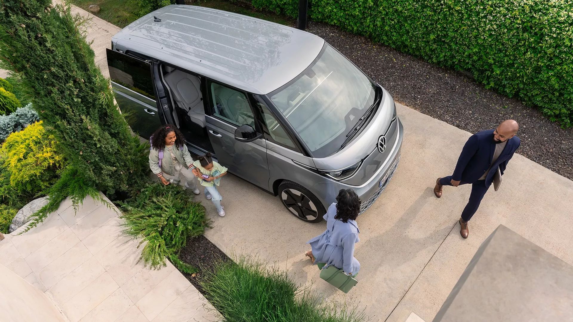 Поглед отгоре към електрическия Volkswagen ID. Buzz пред модерен дом с хора, които се подготвят за излизане