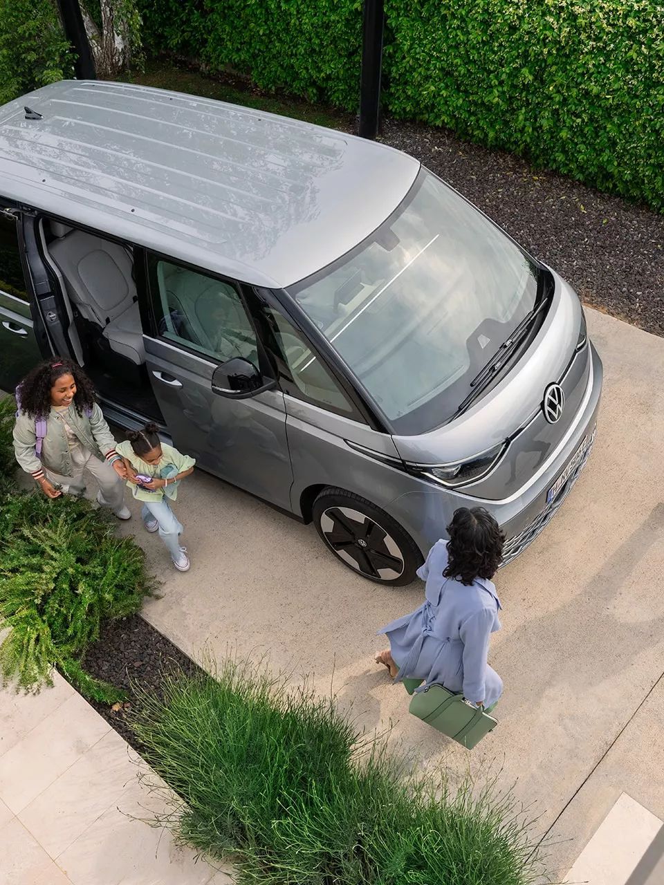 Поглед отгоре към електрическия Volkswagen ID. Buzz пред модерен дом с хора, които се подготвят за излизане