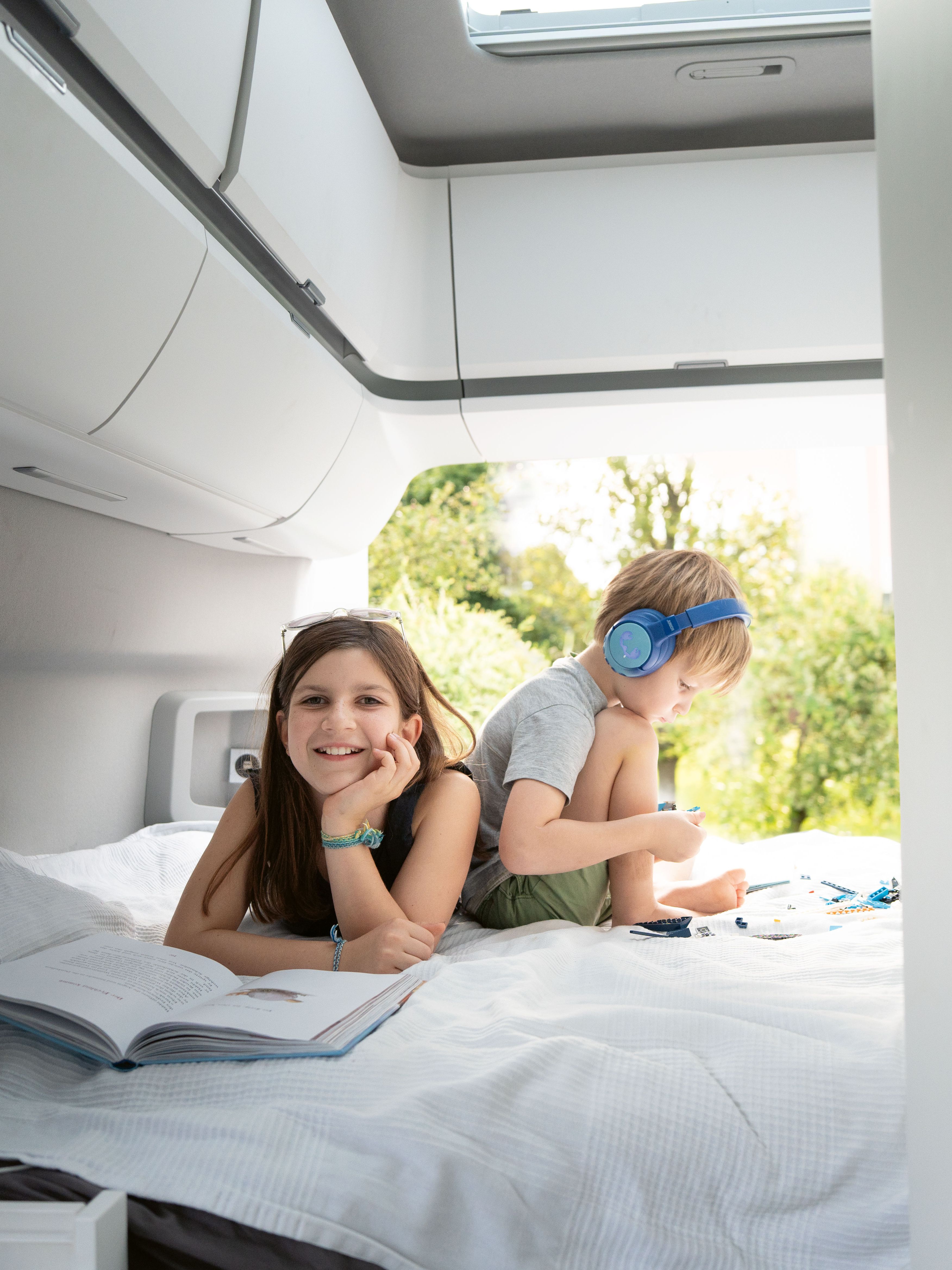 Ein Kind mit Kopfhörern sitzt auf einem Bett und spielt mit einem Tablet, während ein Erwachsener daneben liegt und ein Buch liest. Beide befinden sich in einem hellen Raum mit großen Fenstern.