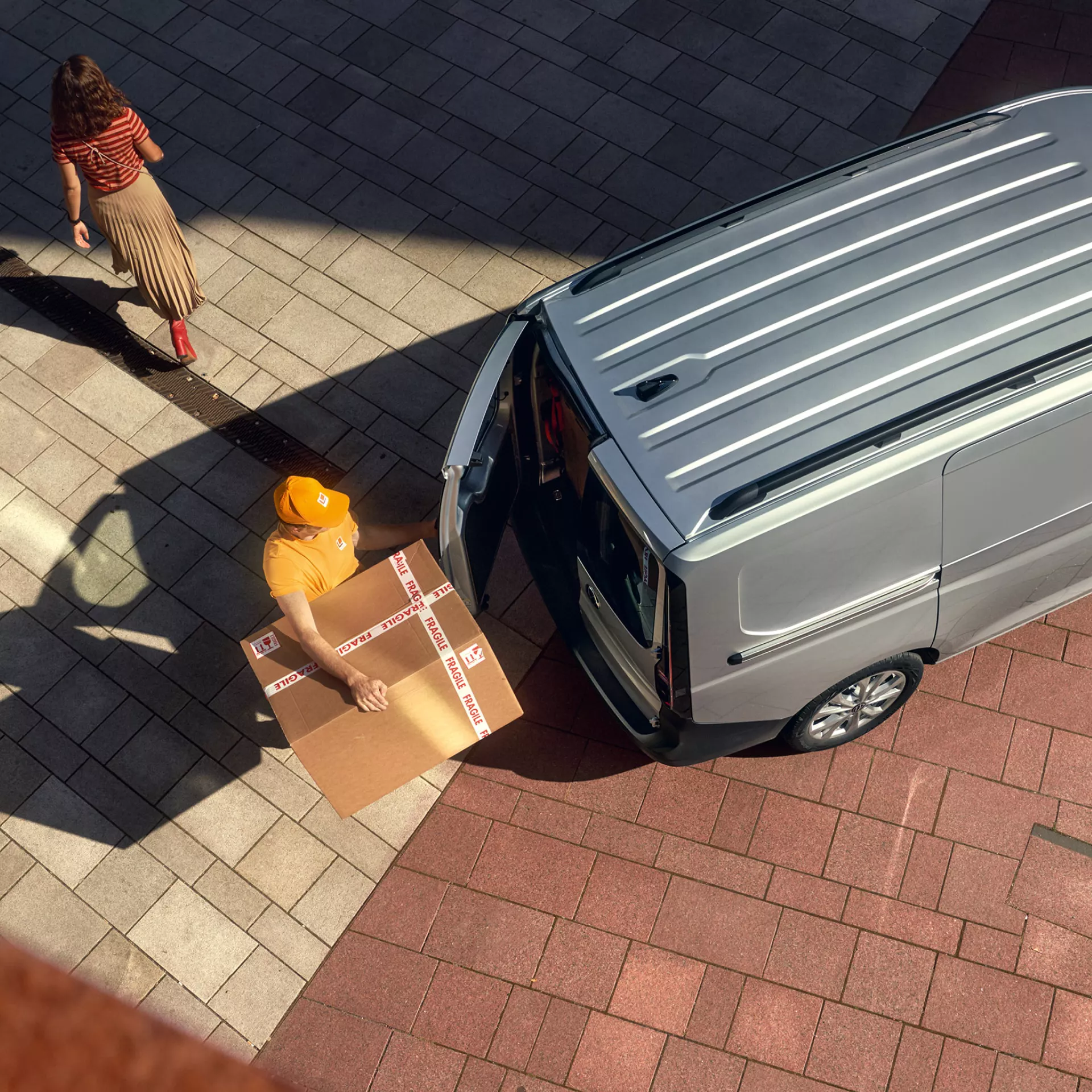 Изглед отгоре на куриер, който товари голям кашон с надпис „чупливо“ в задната част на сребрист товарен ван Volkswagen, докато жена преминава наблизо.