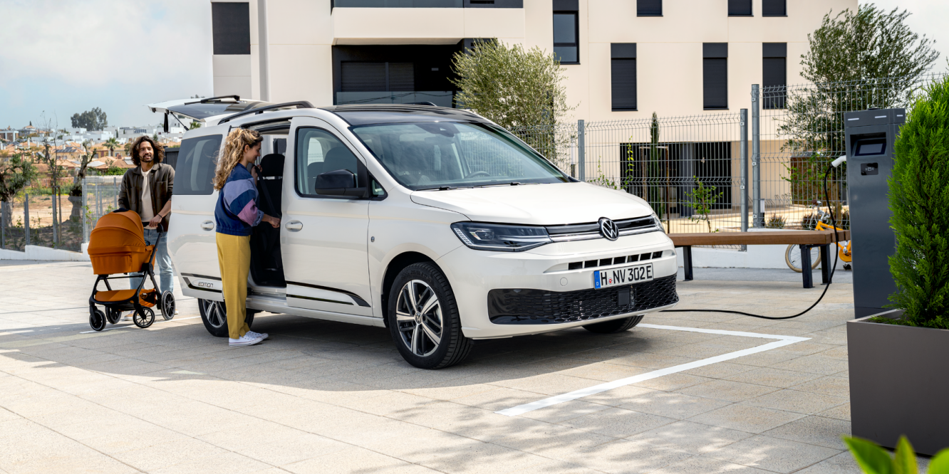 Семейство с детска количка зарежда бял електрически Volkswagen Caddy на зарядна станция пред жилищна сграда