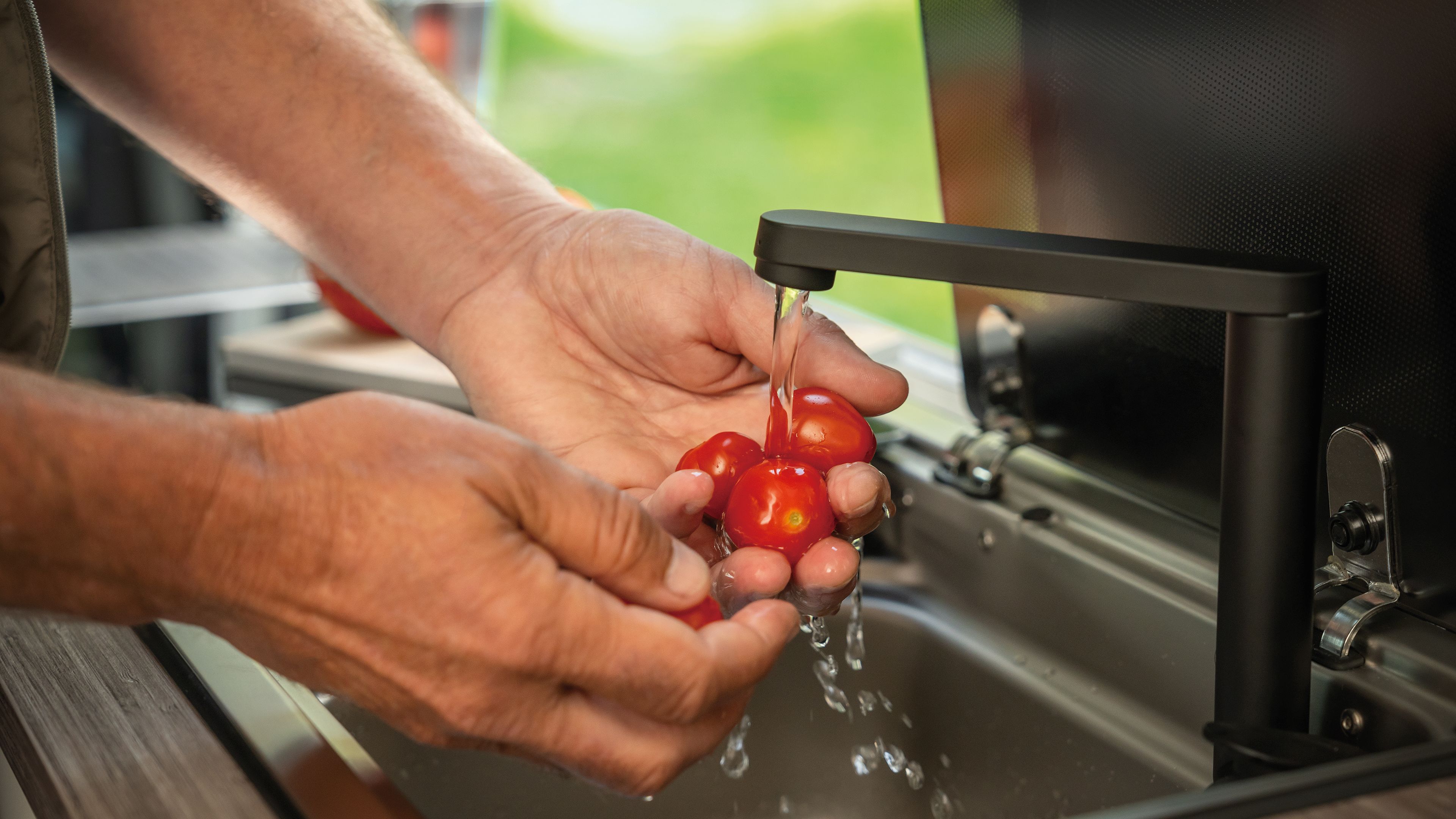 Eine Person wäscht mehrere kleine rote Tomaten unter einem Wasserhahn in einer Küche. Die Hände der Person sind im Fokus und die Tomaten werden gründlich abgespült. Der Hintergrund zeigt ein Fenster mit grünem Außenbereich.
