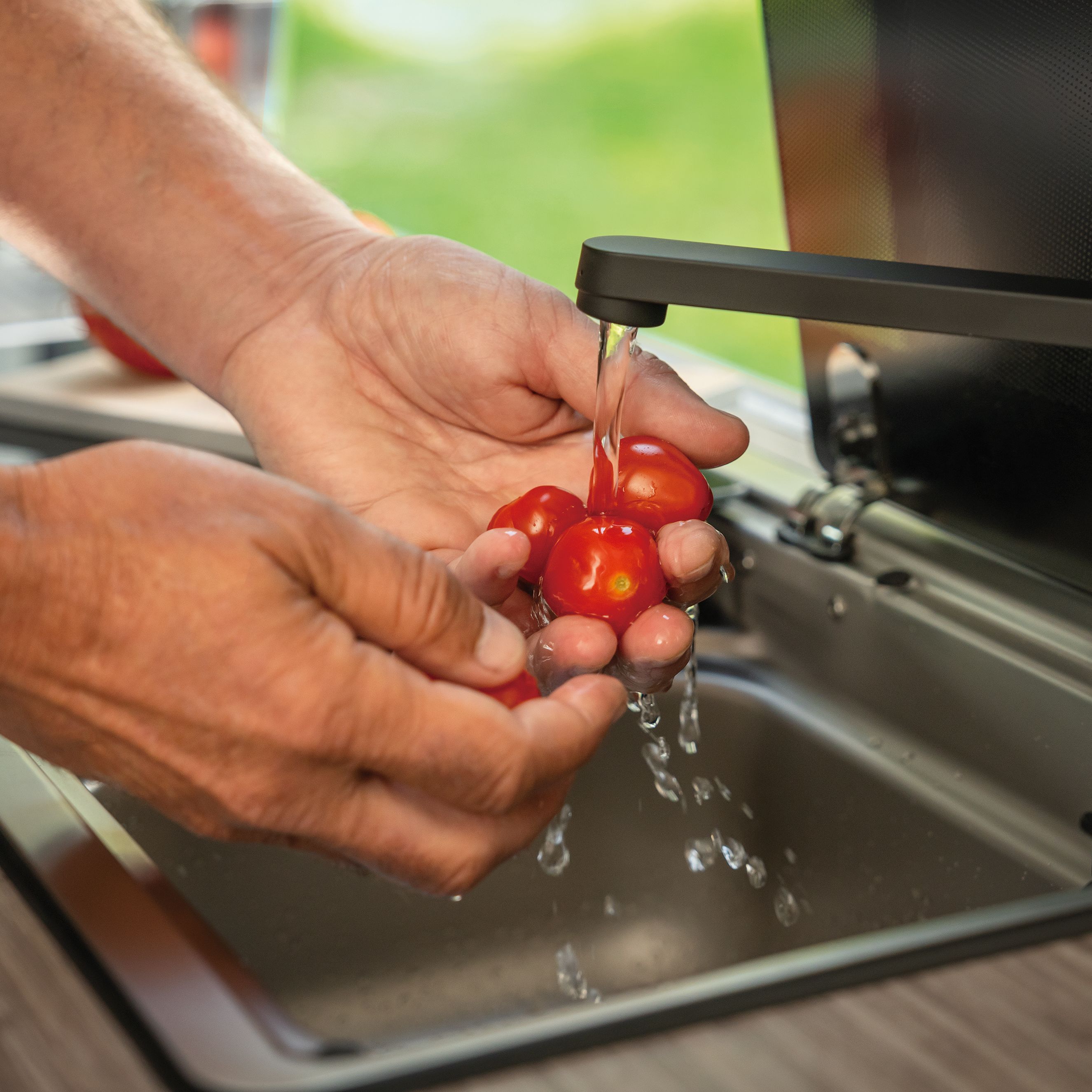 Eine Person wäscht mehrere kleine rote Tomaten unter einem Wasserhahn in einer Küche. Die Hände der Person sind im Fokus und die Tomaten werden gründlich abgespült. Der Hintergrund zeigt ein Fenster mit grünem Außenbereich.
