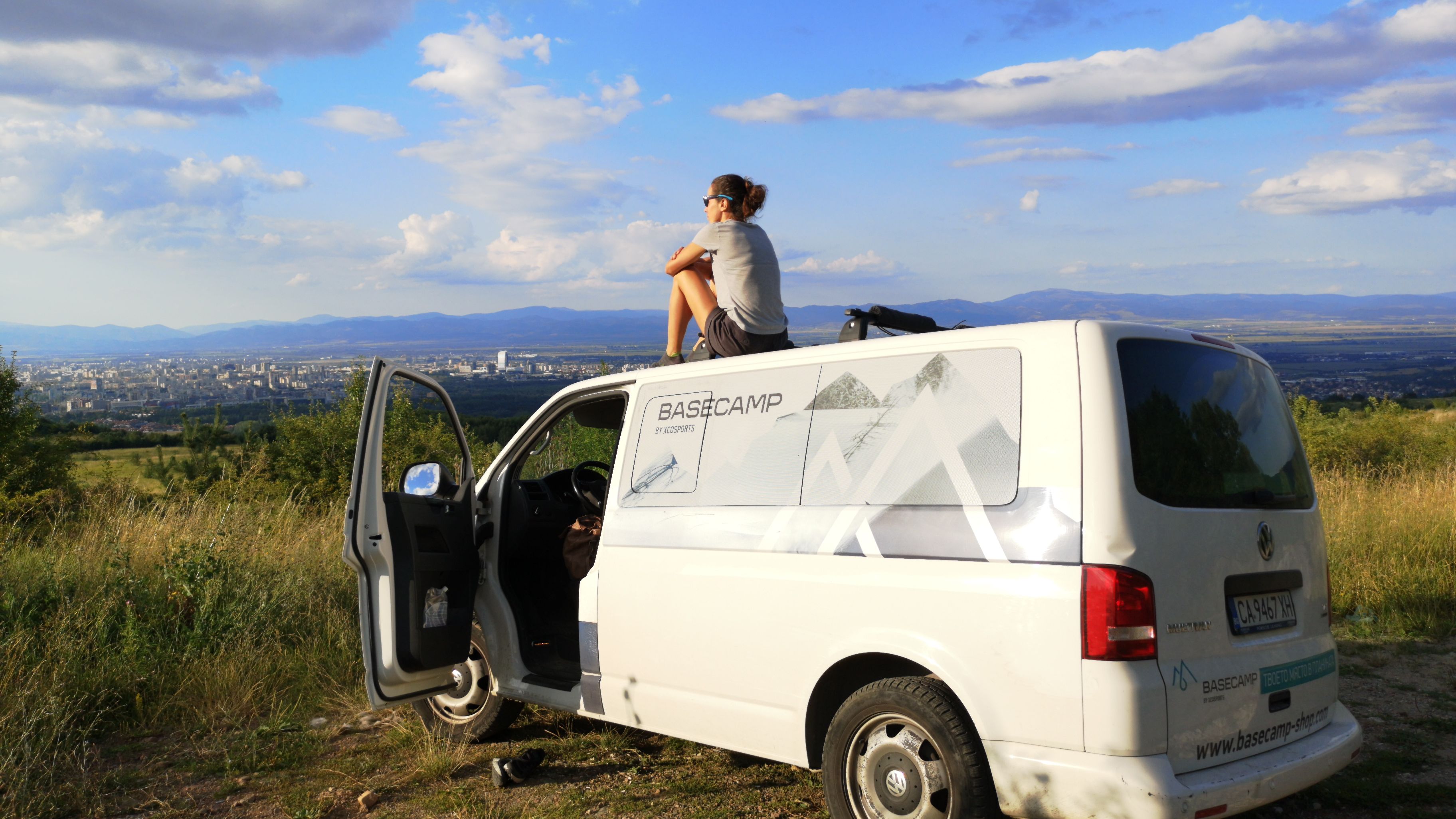 Жена седи върху покрива на бял ван с отворена предна врата, наслаждавайки се на красивата гледка. В далечината се вижда град с множество сгради, заобиколен от хълмове и облачно небе.