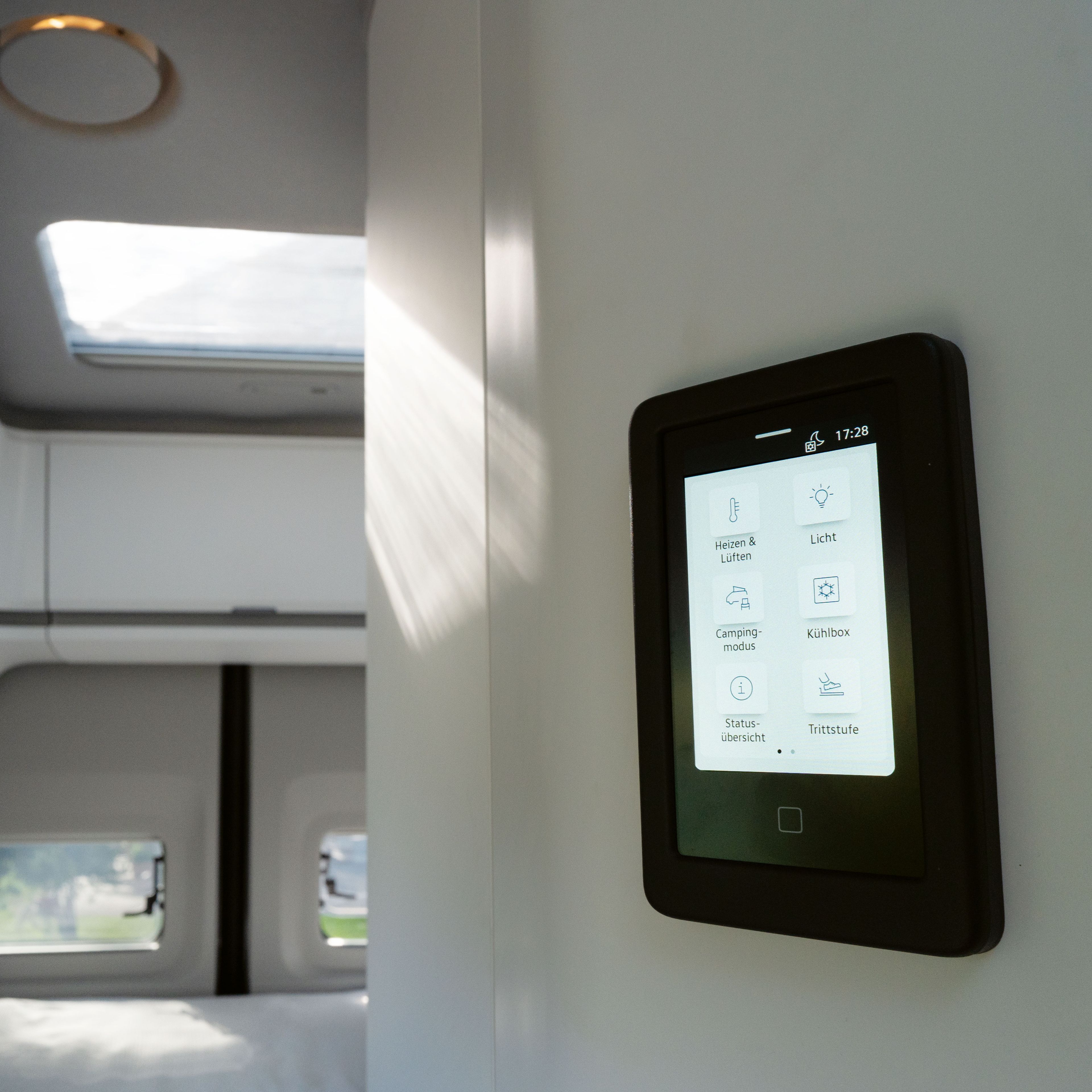 Ein modernes Wohnmobilinterieur mit einem Touchscreen-Bedienfeld an der Wand. Das Bedienfeld zeigt verschiedene Steueroptionen für Beleuchtung, Klima und andere Funktionen. Im Hintergrund sind Betten und Fenster sichtbar.