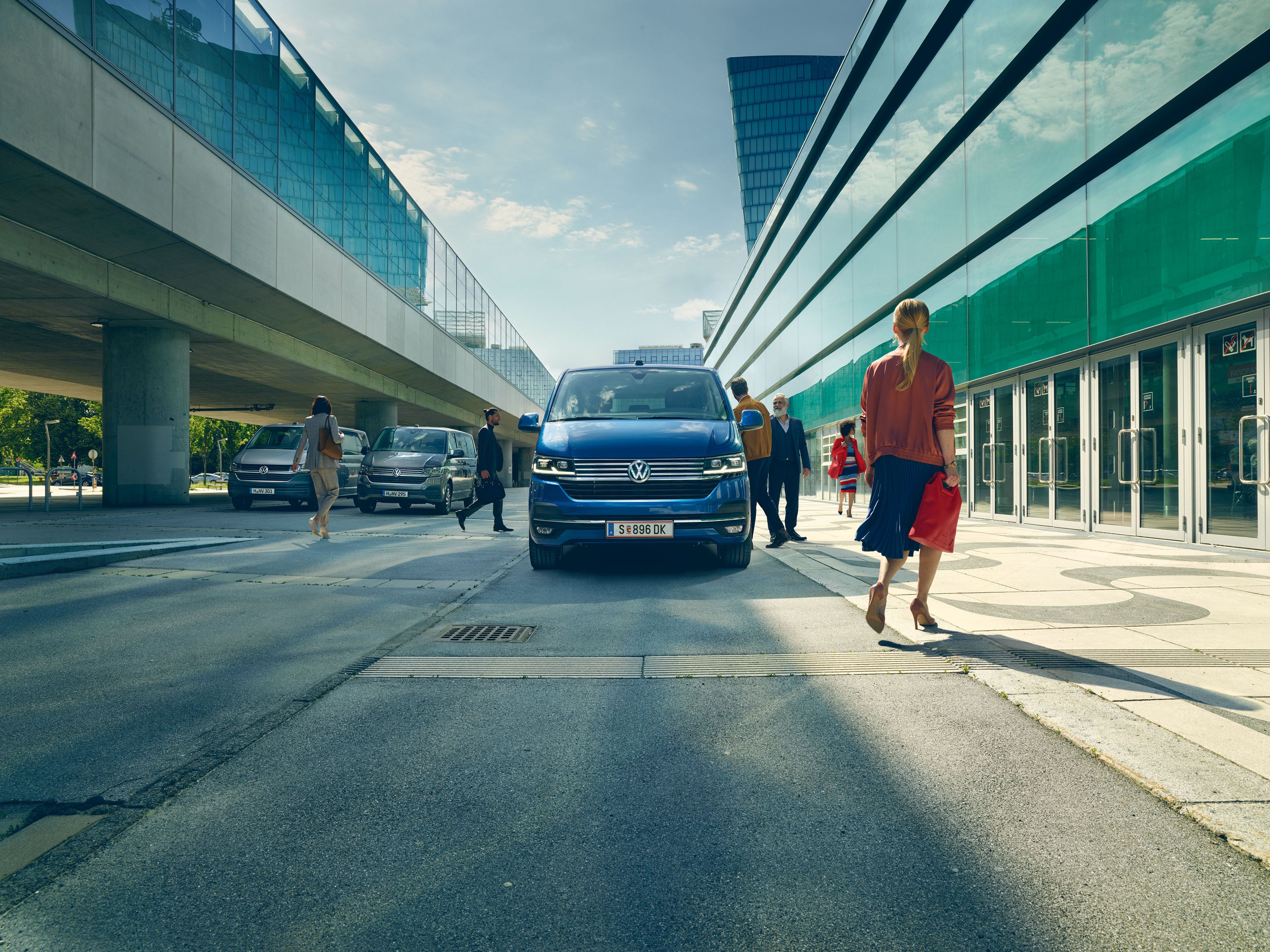 Blauer VW Caravelle t6 steht am Straßenrand und ist von vorne zu sehen. Passanten gehen daran vorbei. 
