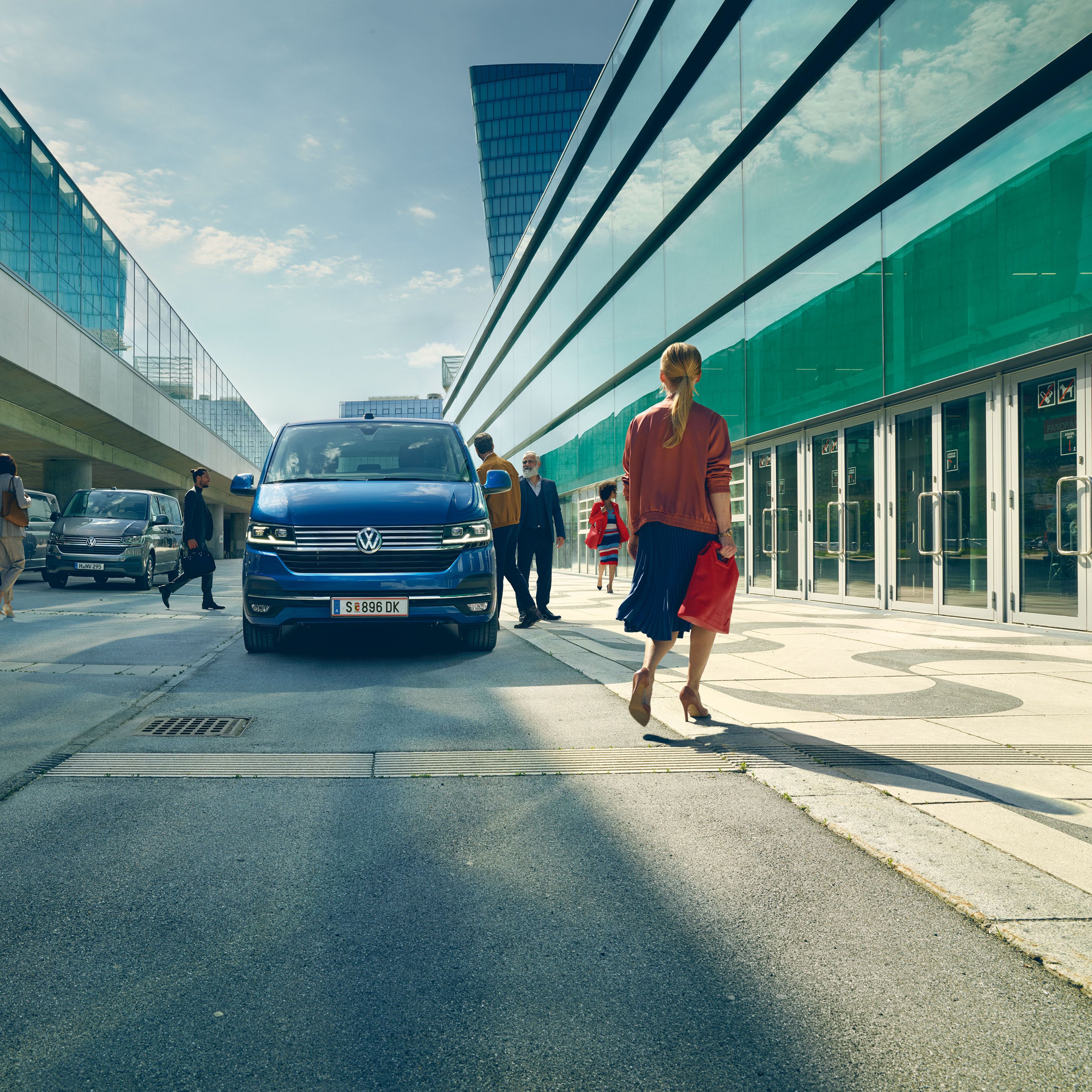 Blauer VW Caravelle t6 steht am Straßenrand und ist von vorne zu sehen. Passanten gehen daran vorbei. 