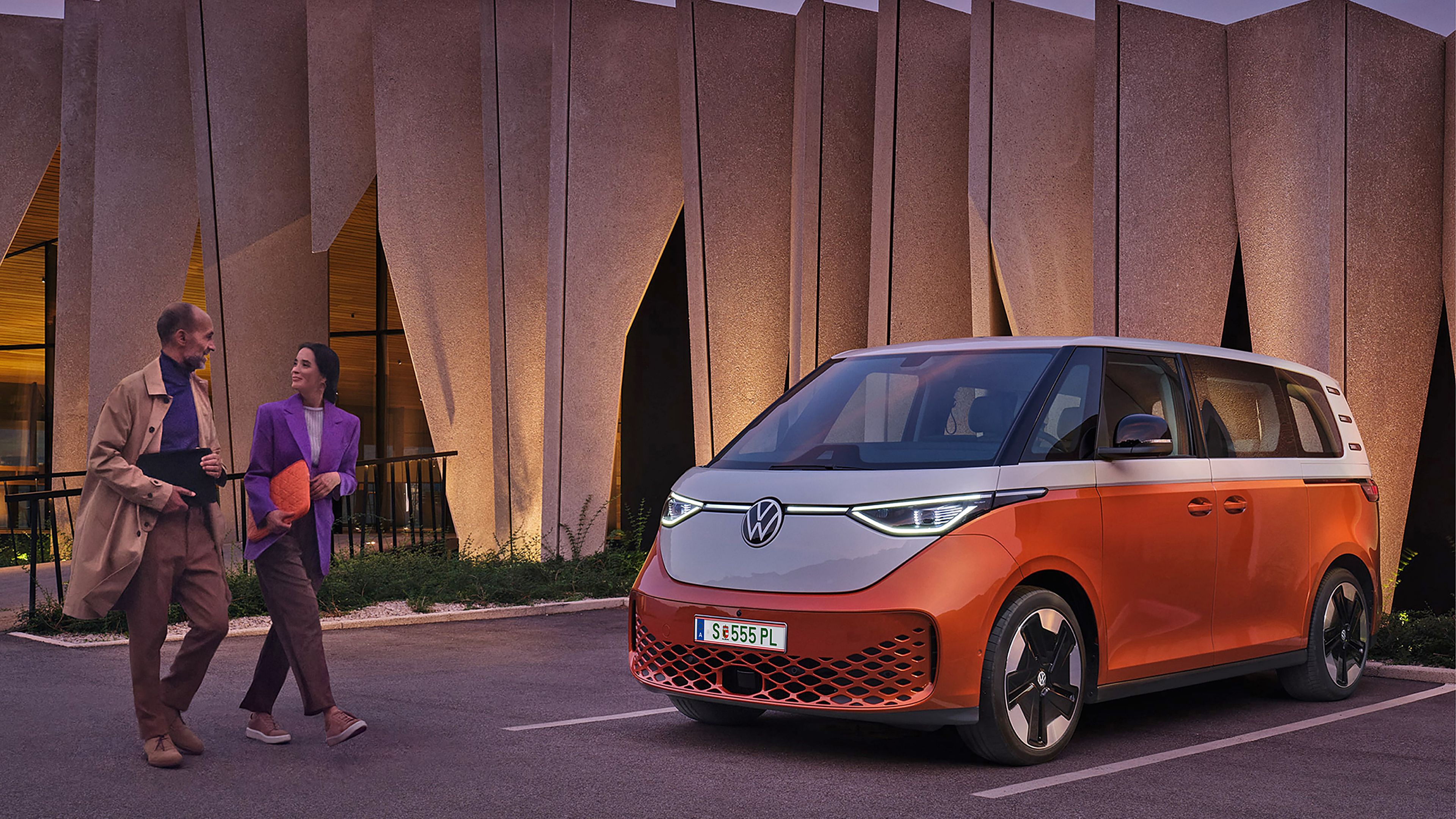 Двойка пред модерен сграден силует с паркиран електрически Volkswagen ID. Buzz