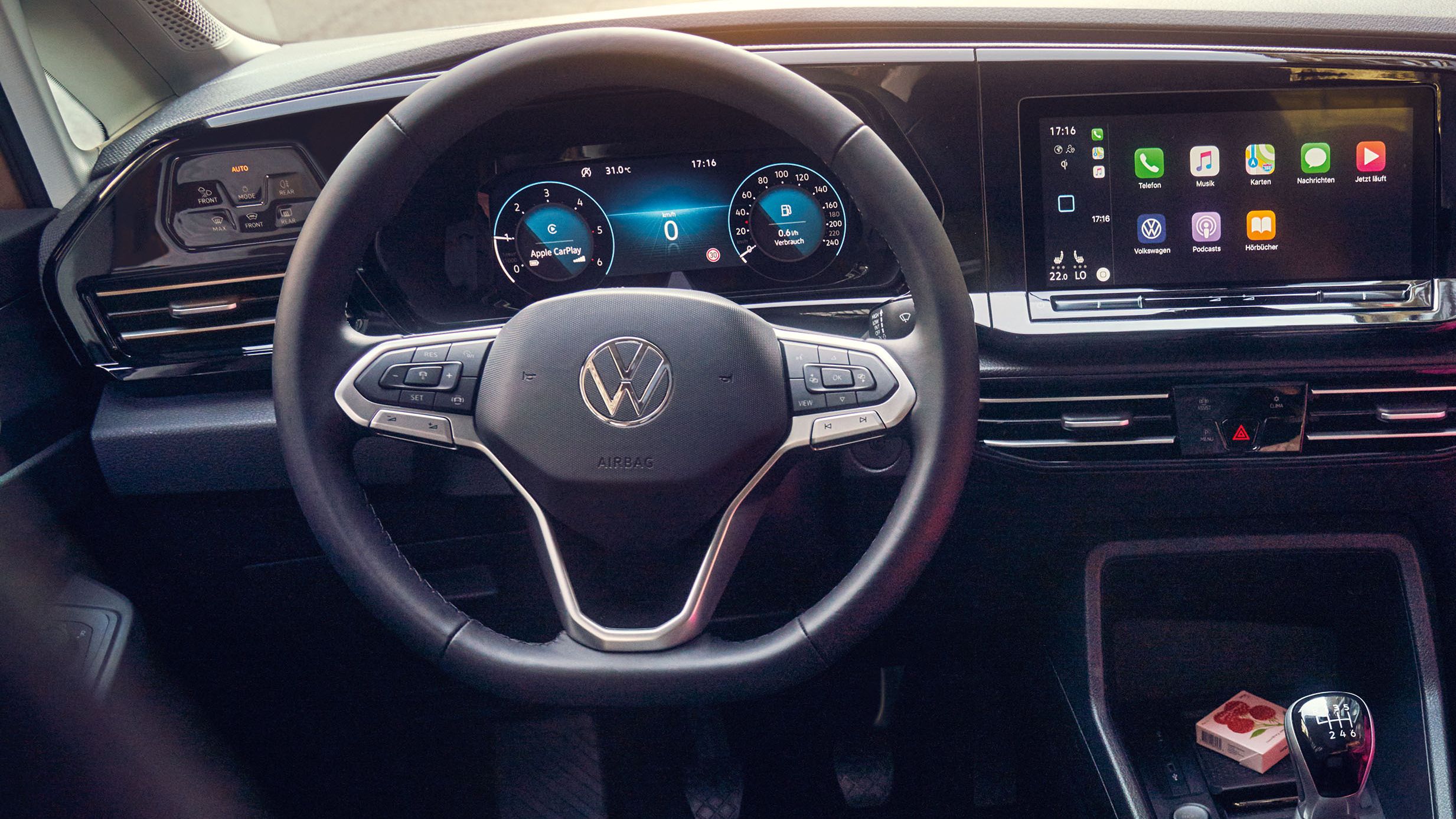 Интериор на автомобил Volkswagen с черни кожени седалки, ръчно скоростен лост и мултимедиен дисплей. Женска ръка с гривна и розова пола се вижда на предната пътническа седалка.