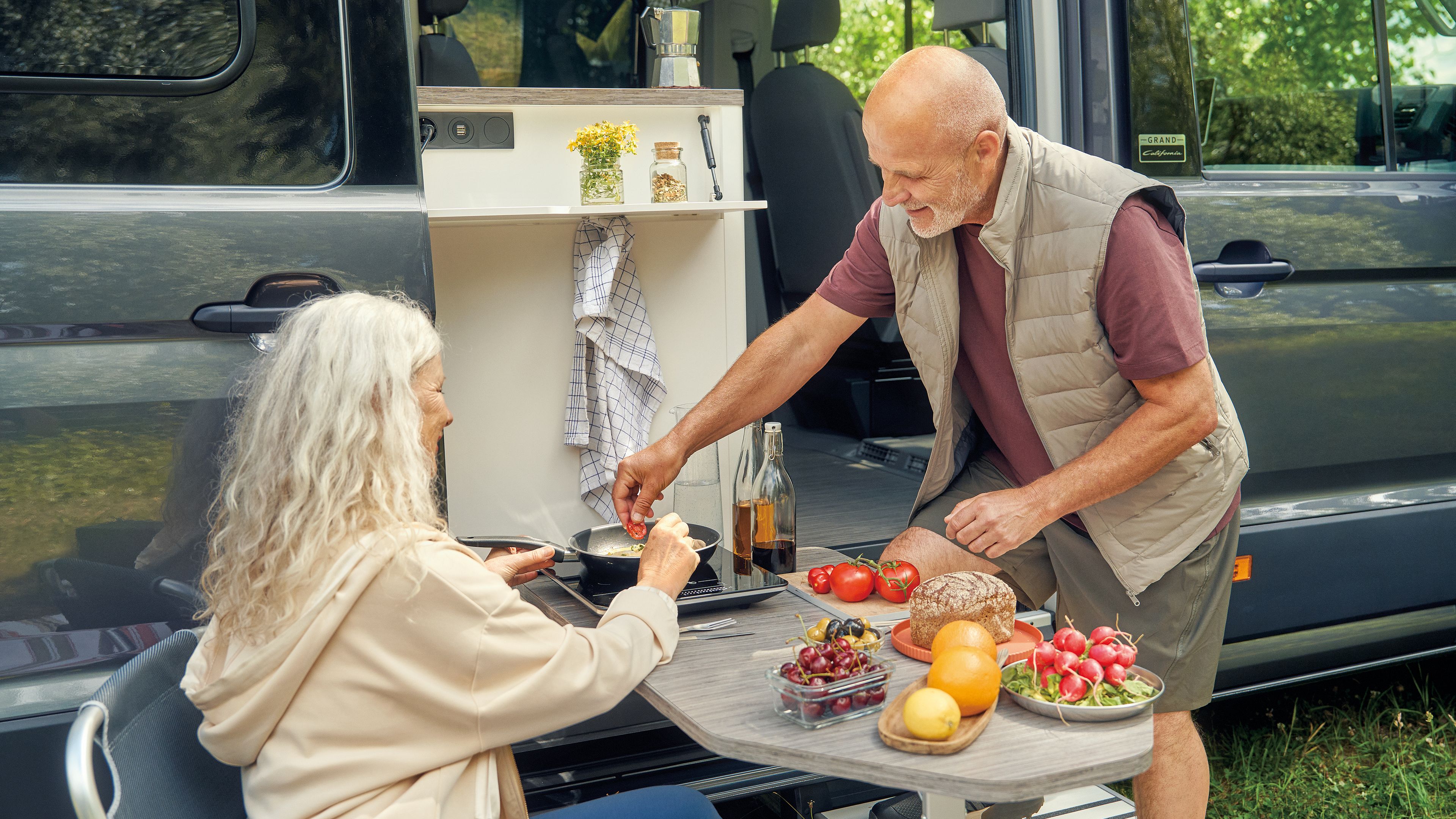 Ein älteres Paar sitzt vor einem Wohnmobil und bereitet gemeinsam eine Mahlzeit zu. Auf dem Tisch befinden sich verschiedene frische Lebensmittel, darunter Tomaten, Radieschen und Brot. Die Umgebung ist grün und einladend.