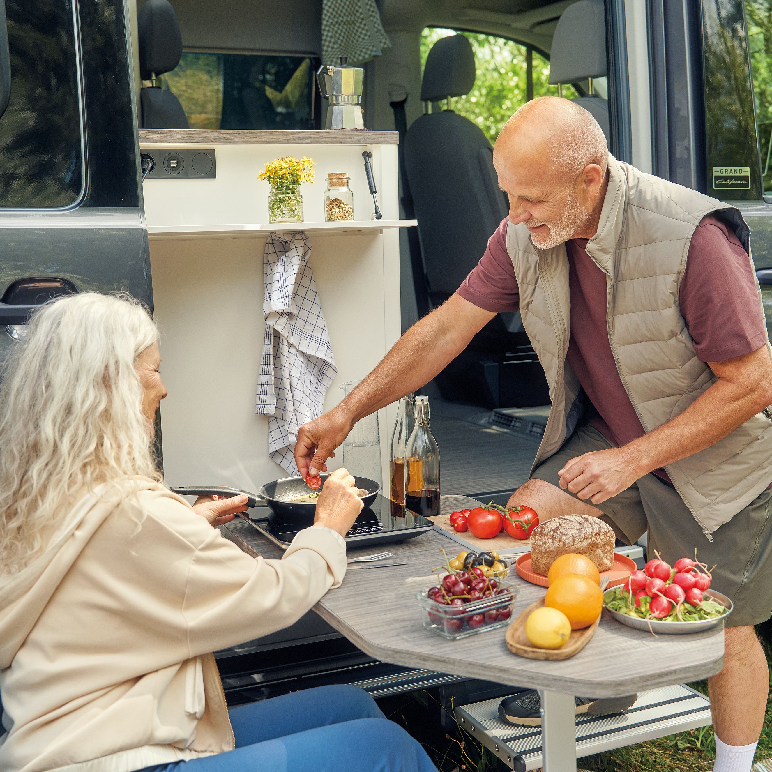 Ein älteres Paar sitzt vor einem Wohnmobil und bereitet gemeinsam eine Mahlzeit zu. Auf dem Tisch befinden sich verschiedene frische Lebensmittel, darunter Tomaten, Radieschen und Brot. Die Umgebung ist grün und einladend.
