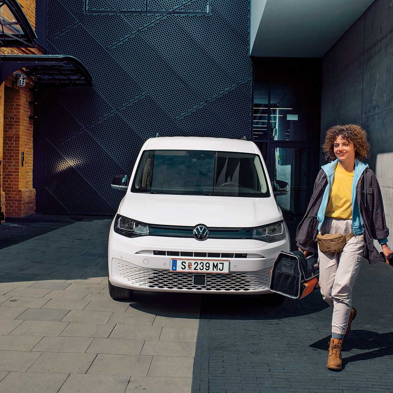 Усмихната жена в работно облекло носи инструменти, минавайки покрай бял Volkswagen Caddy, паркиран пред модерен индустриален комплекс.