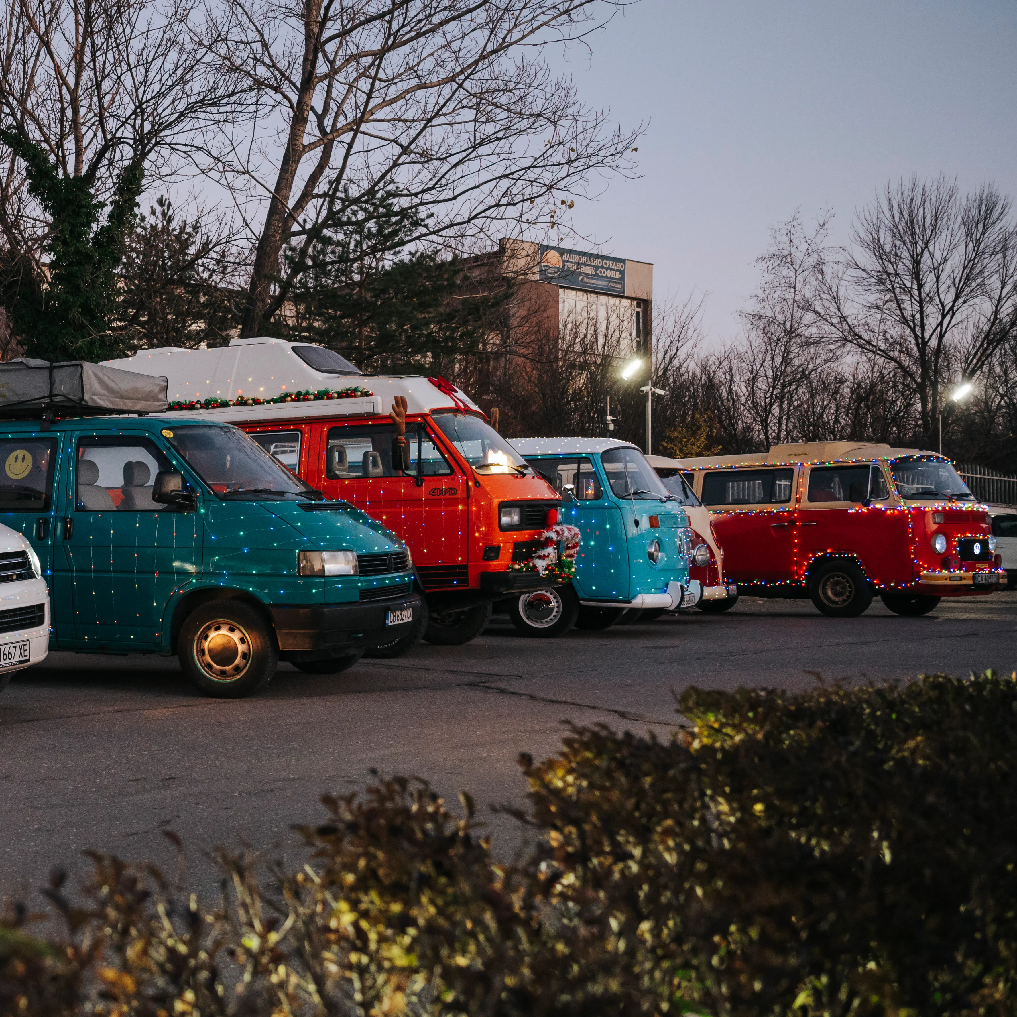 Редица кемпери и микробуси Volkswagen са паркирани на открит паркинг по здрач. На преден план има растения, а отзад са видими голи дървета и сграда. Някои от превозните средства са ярко оцветени и имат различни дизайни.