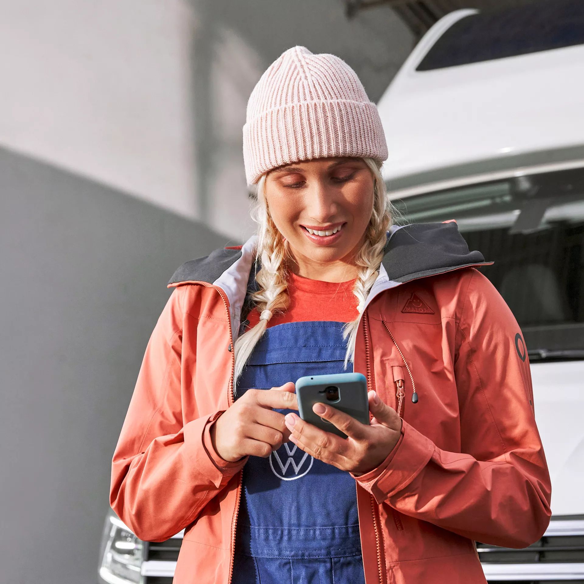 Eine Frau mit blonden Zöpfen und Mütze steht vor einem Wohnmobil und benutzt ein Smartphone. Sie trägt eine rote Jacke und blaue Arbeitshose. Ihr Gesicht ist verschwommen.