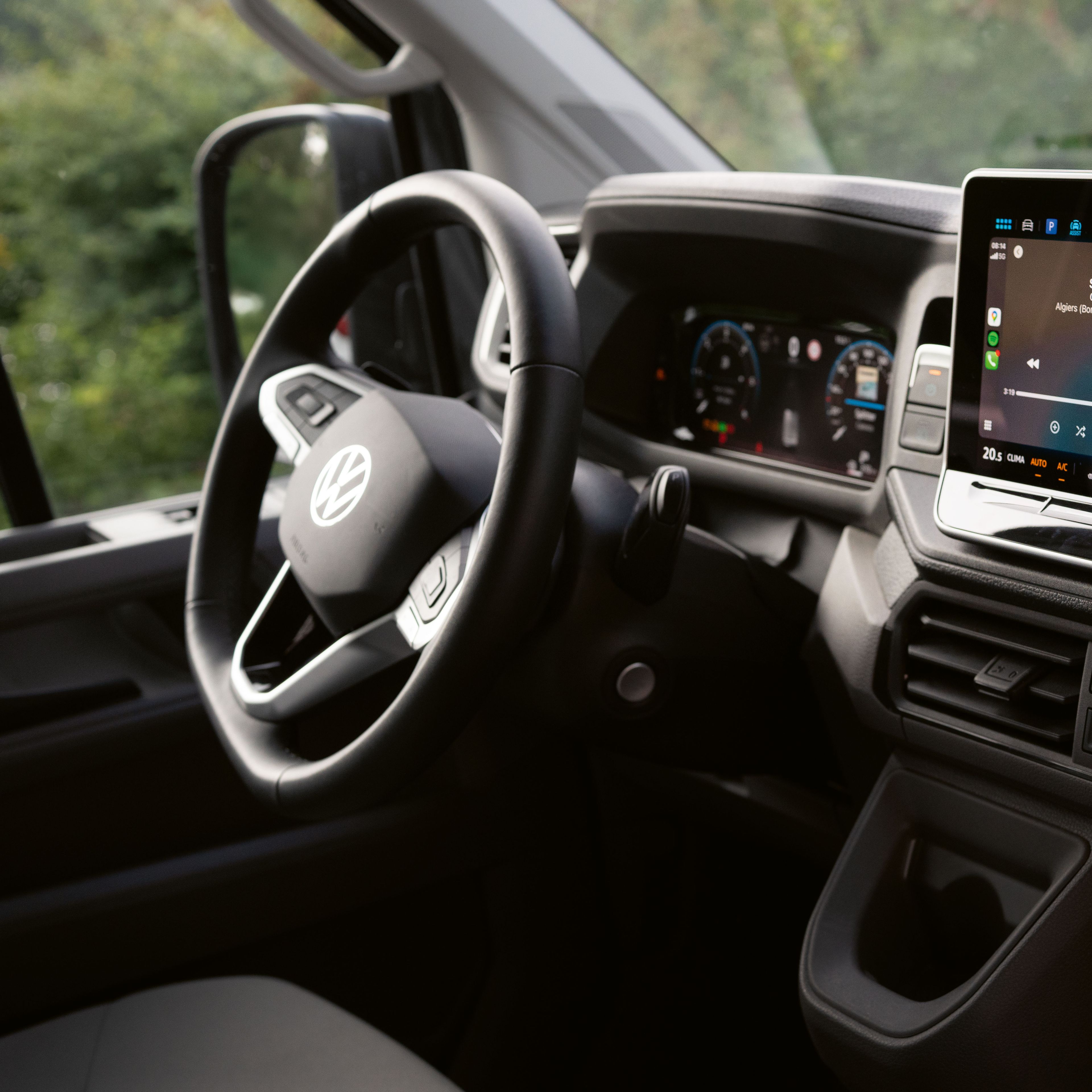 Das Bild zeigt das Innere eines Volkswagen-Autos, genauer gesagt das Armaturenbrett und das Lenkrad. Auf dem Armaturenbrett befindet sich ein großer Touchscreen, der Multimedia-Inhalte anzeigt. Im Hintergrund sind grüne Bäume zu sehen.