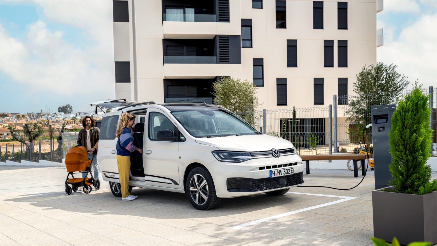 Семейство с детска количка зарежда бял електрически Volkswagen Caddy на зарядна станция пред жилищна сграда