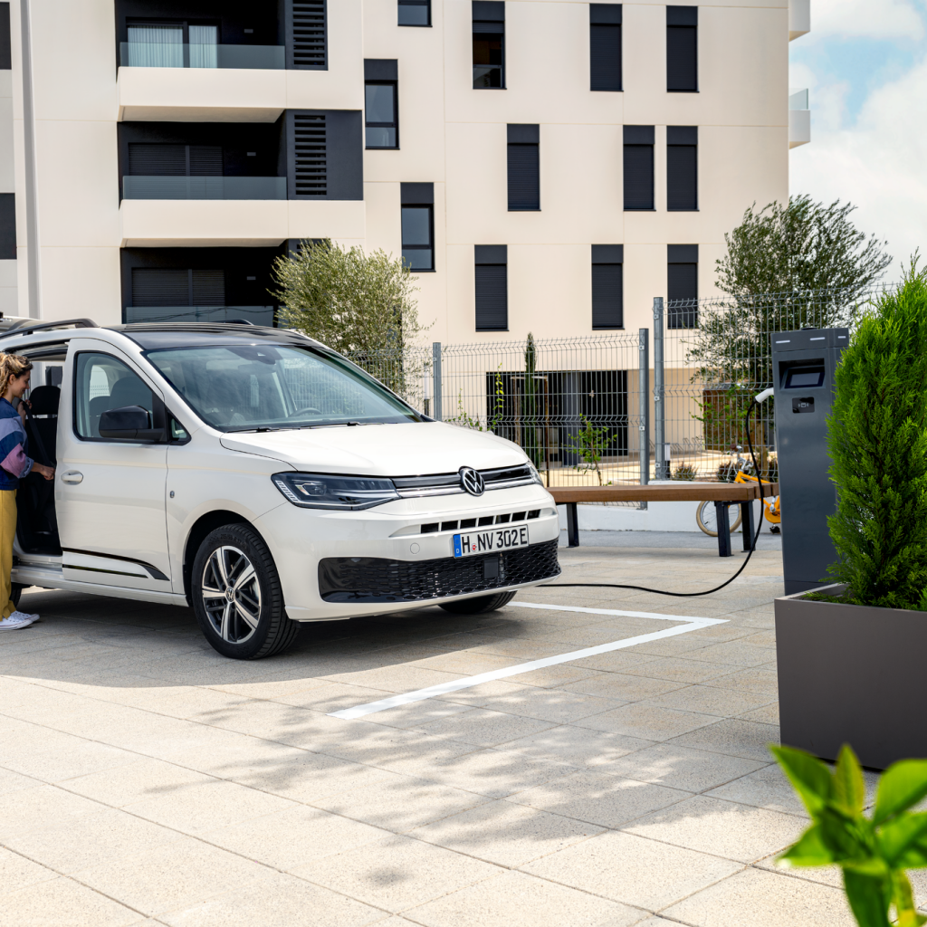 Семейство с детска количка зарежда бял електрически Volkswagen Caddy на зарядна станция пред жилищна сграда