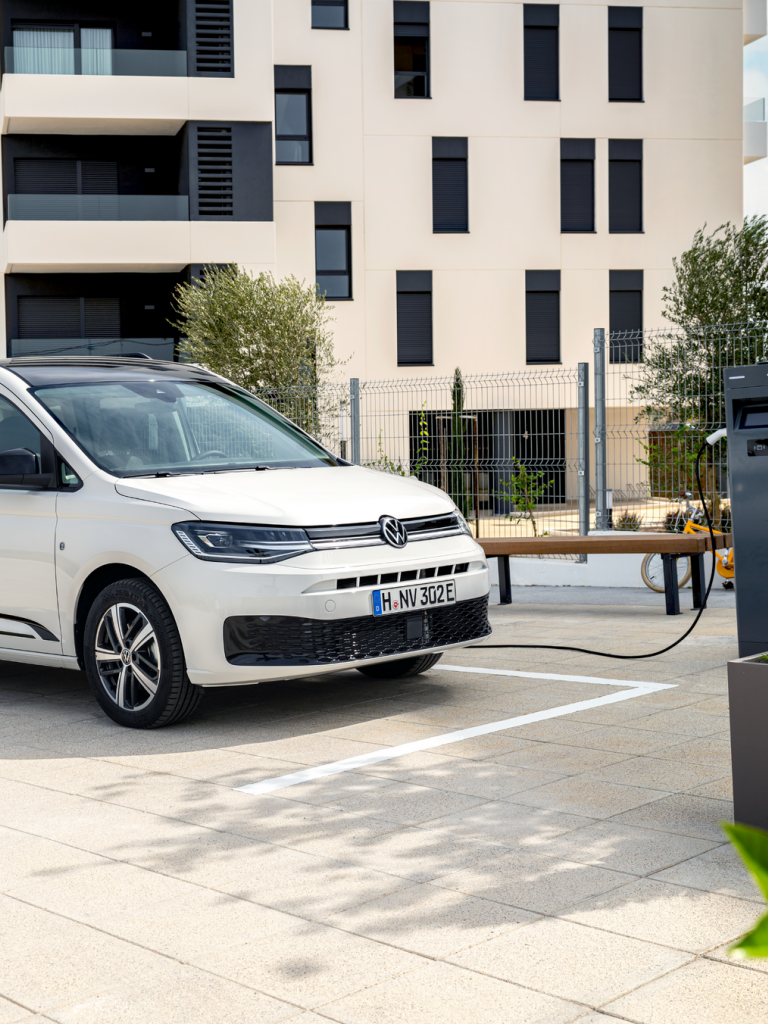 Семейство с детска количка зарежда бял електрически Volkswagen Caddy на зарядна станция пред жилищна сграда