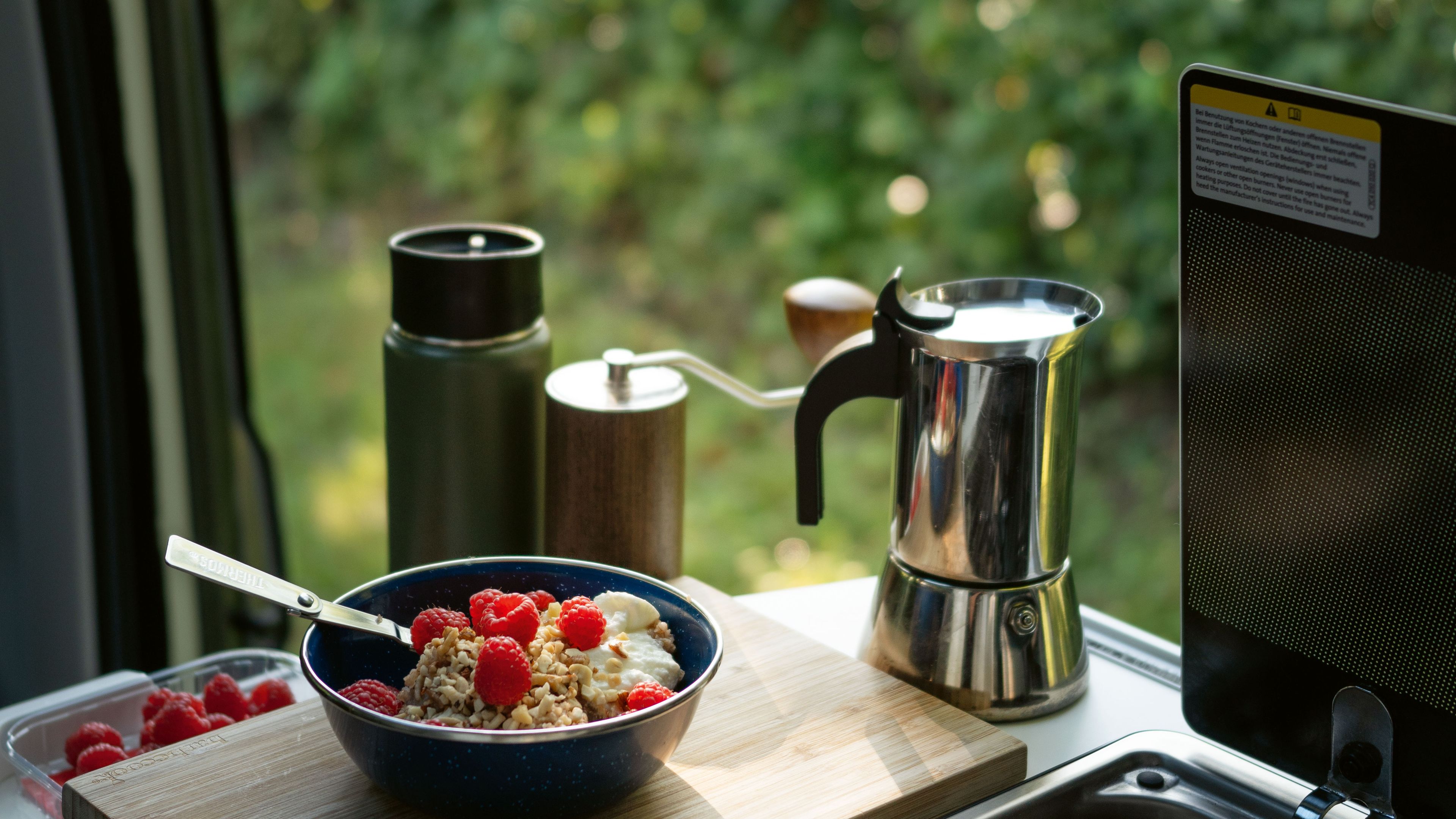 Eine Schüssel mit Müsli, frischen Himbeeren und einem Löffel steht auf einem Schneidebrett in einer Campingküche. Daneben befinden sich eine Espressokanne, eine Kaffeemühle und eine Thermoskanne. Im Hintergrund ist durch das Fenster eine grüne Landschaft sichtbar.