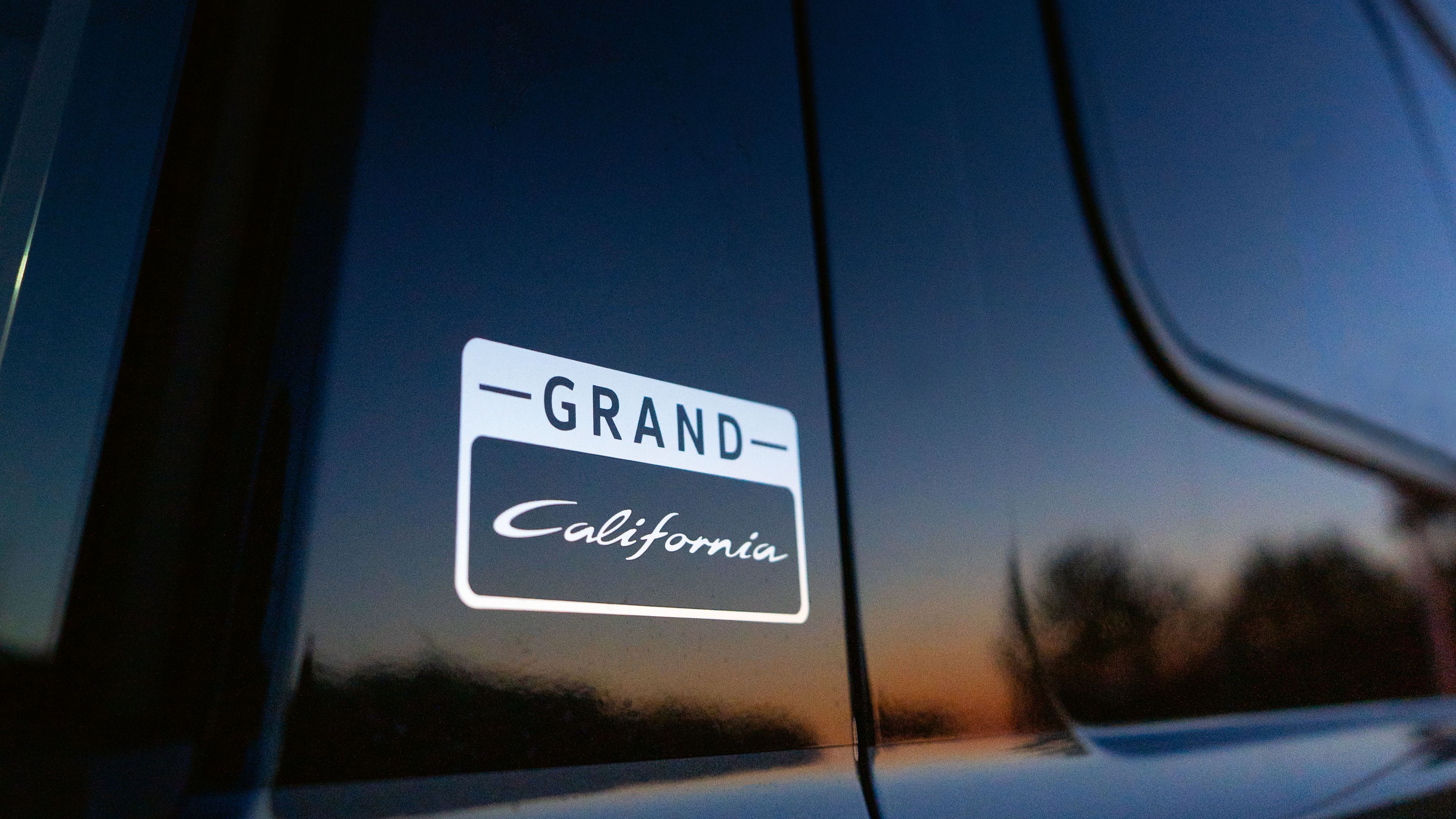Nahaufnahme eines schwarzen Autos mit einem "Grand California" Emblem auf der Seite. Der Sonnenuntergang spiegelt sich im Fenster.