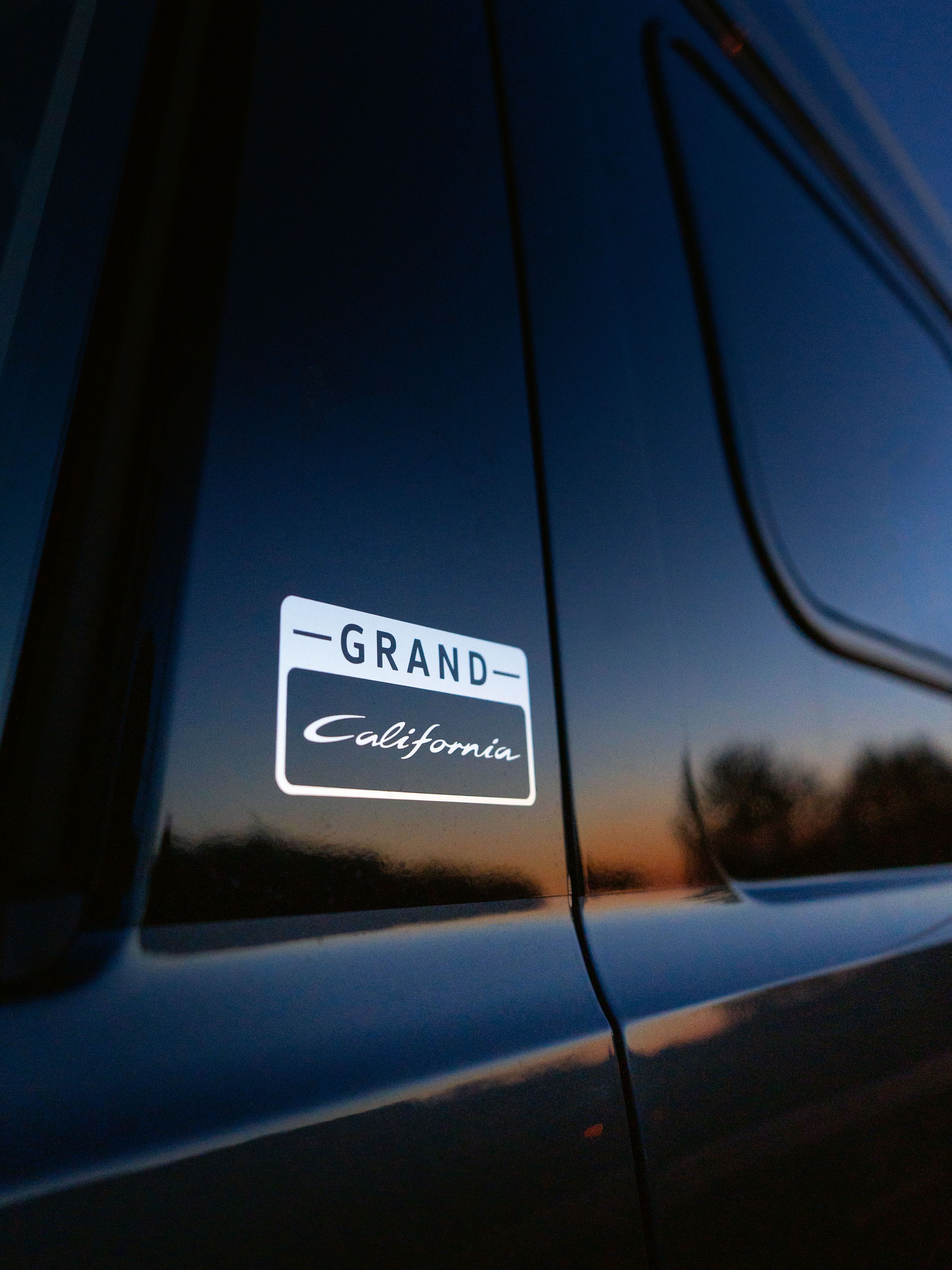 Nahaufnahme eines schwarzen Autos mit einem "Grand California" Emblem auf der Seite. Der Sonnenuntergang spiegelt sich im Fenster.
