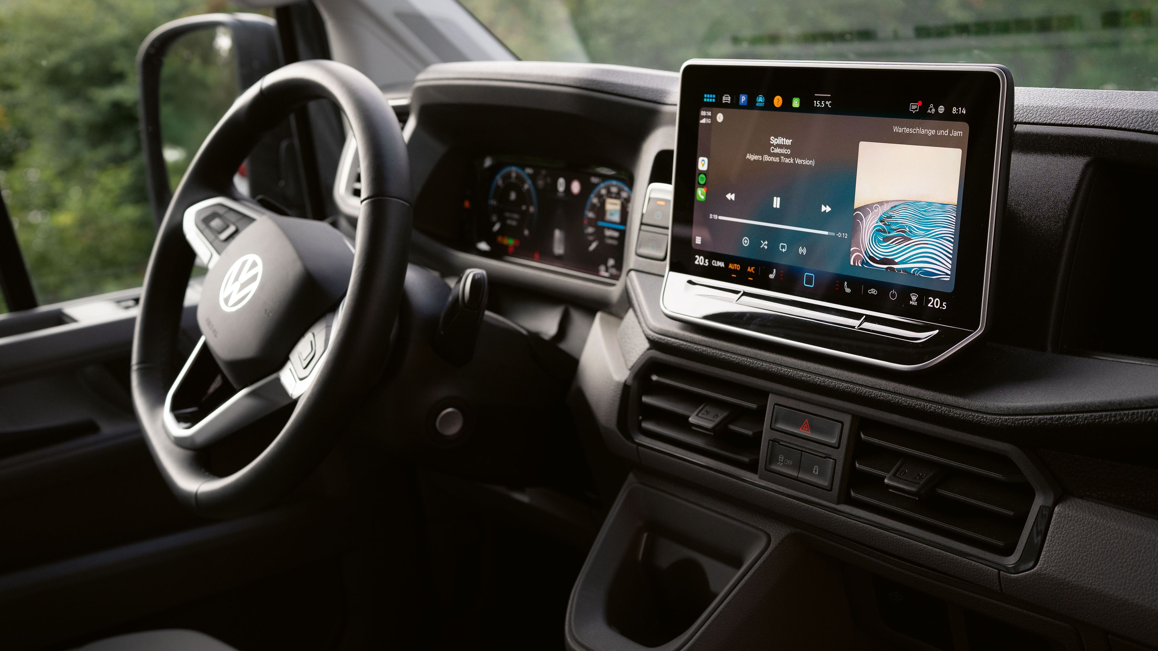 Das Bild zeigt das Innere eines Volkswagen-Autos, genauer gesagt das Armaturenbrett und das Lenkrad. Auf dem Armaturenbrett befindet sich ein großer Touchscreen, der Multimedia-Inhalte anzeigt. Im Hintergrund sind grüne Bäume zu sehen.