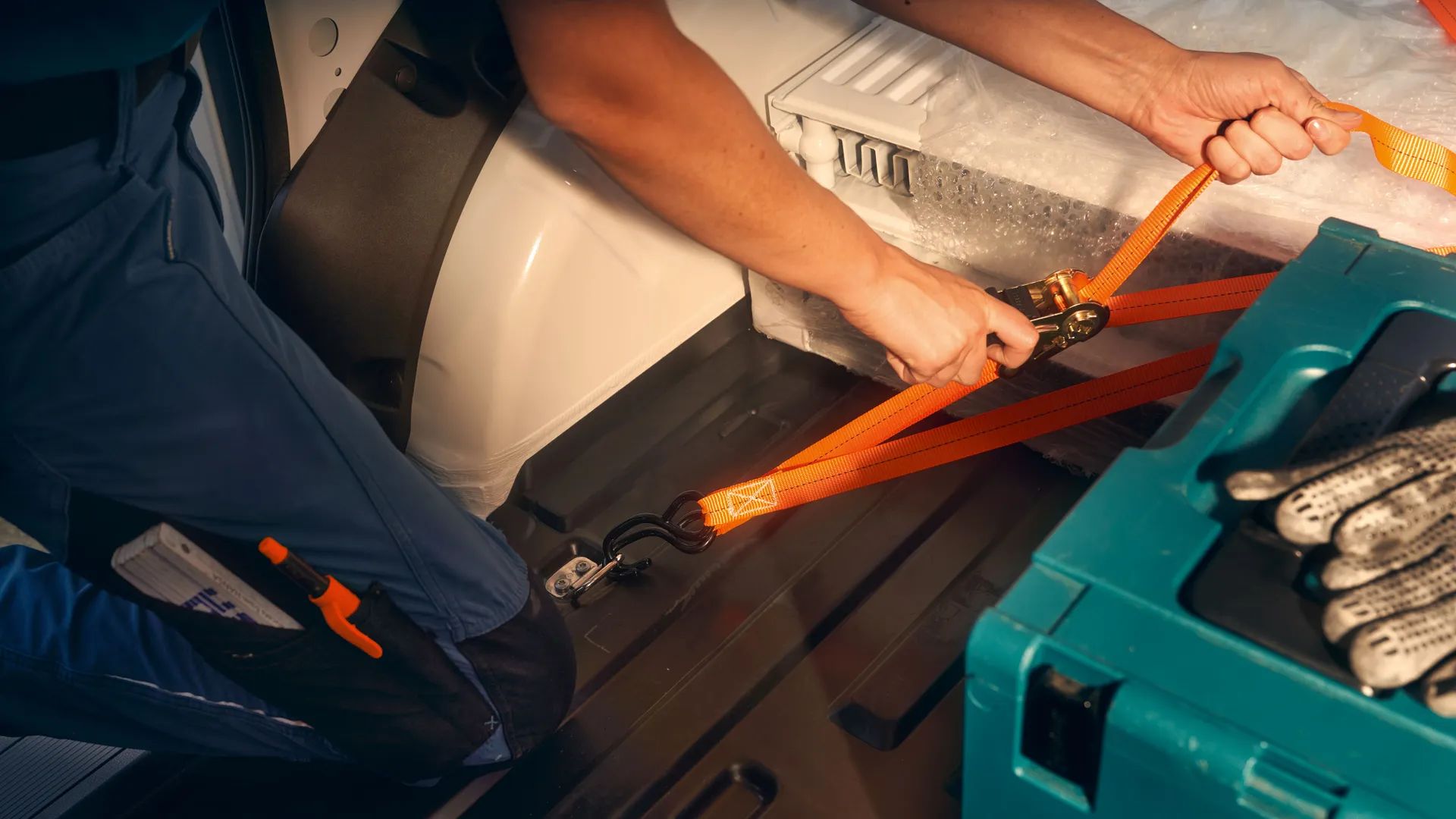 Човек затяга оранжеви ремъци за закрепване на товар в товарно превозно средство. На заден план има голям предмет, покрит с пяна, а отпред - син пластмасов куфар за инструменти. Видими са и чифт защитни ръкавици на седалката.