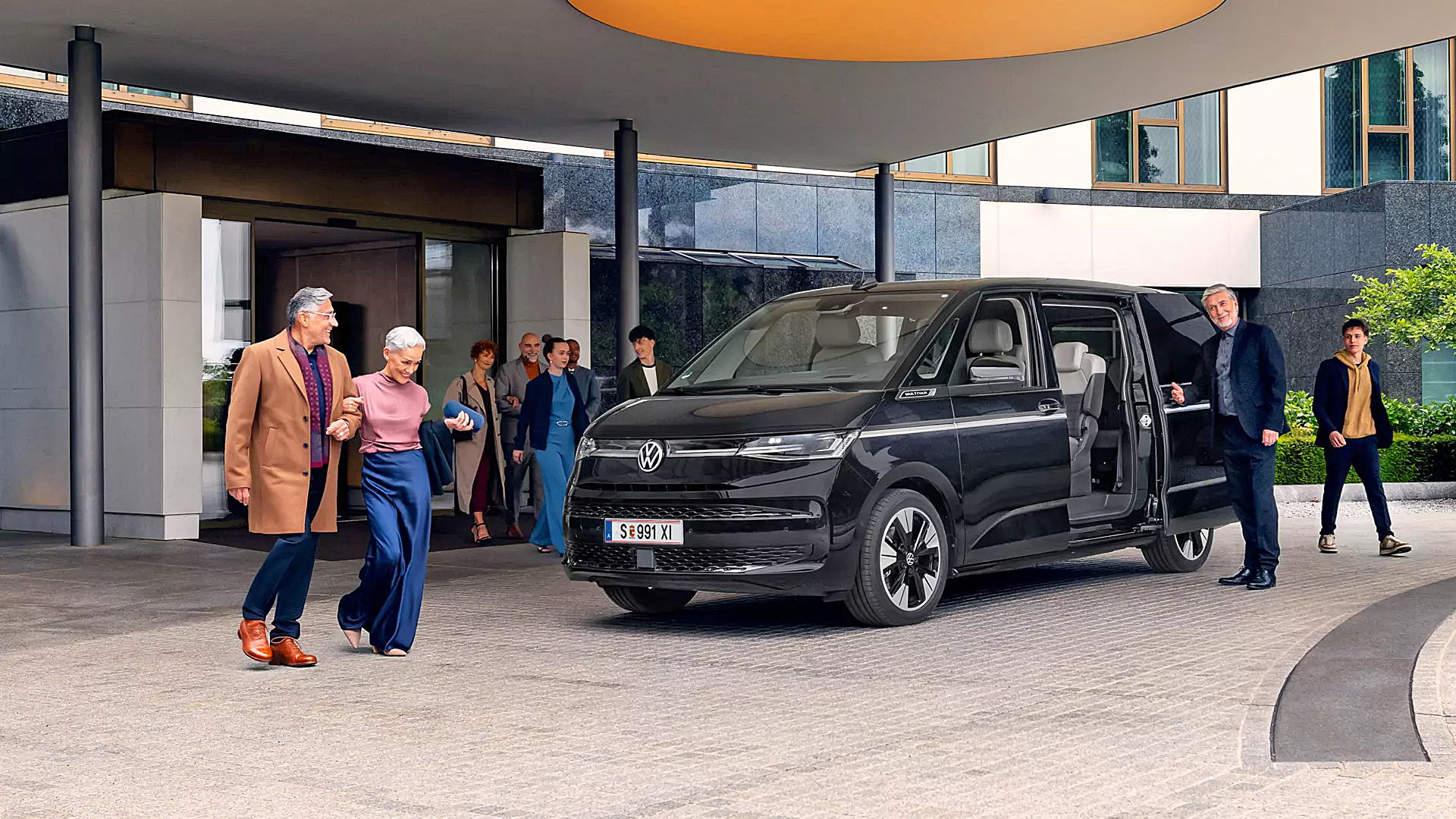 Eine Gruppe von Menschen steht vor einem modernen Gebäude. Ein schwarzer Volkswagen-Bus parkt unter einem überdachten Bereich. Einige Personen steigen aus oder unterhalten sich neben dem Fahrzeug.