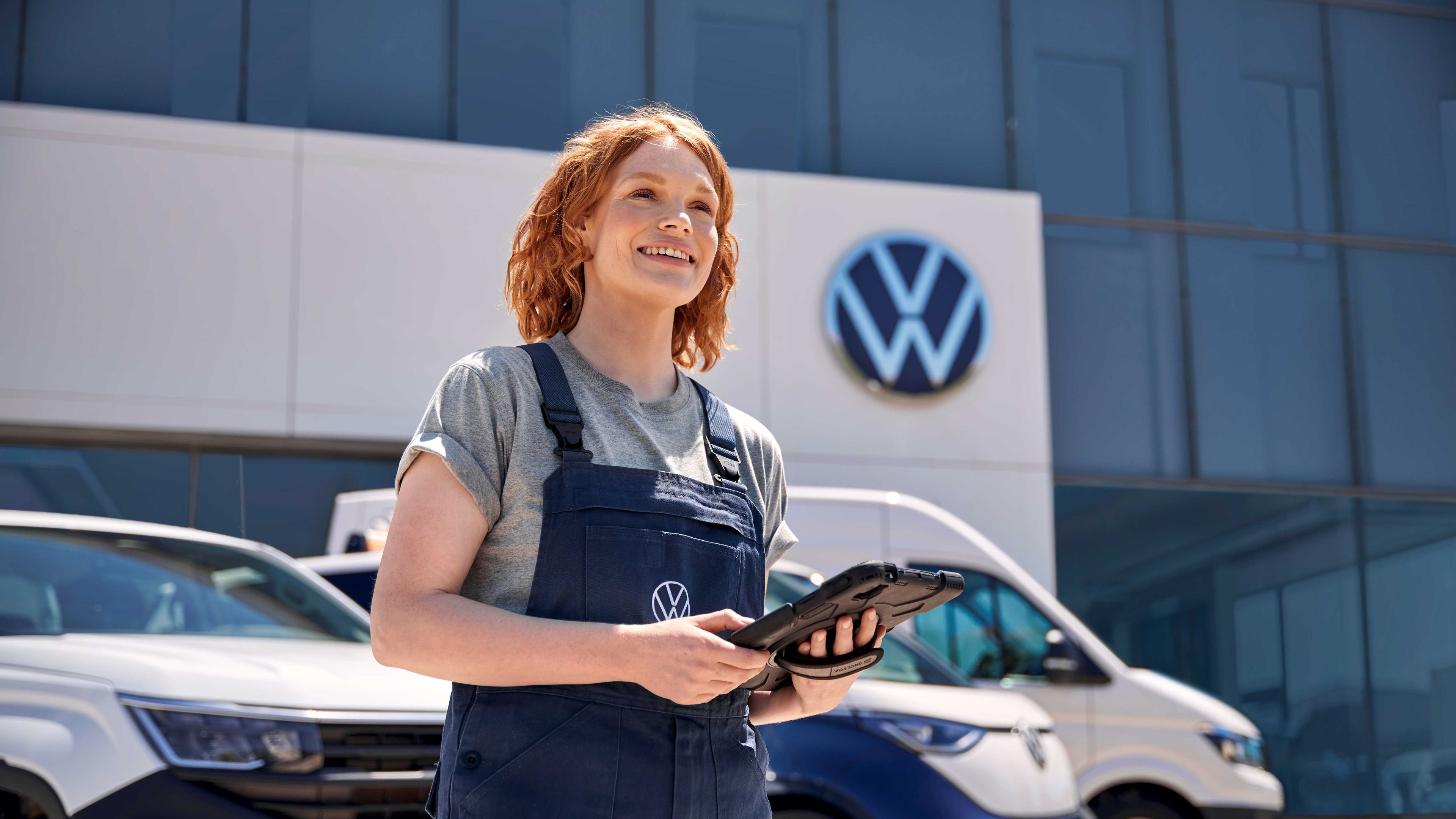 Служител с таблет пред сграда на Volkswagen, с три автомобила на заден план – два бели вана и син автомобил.